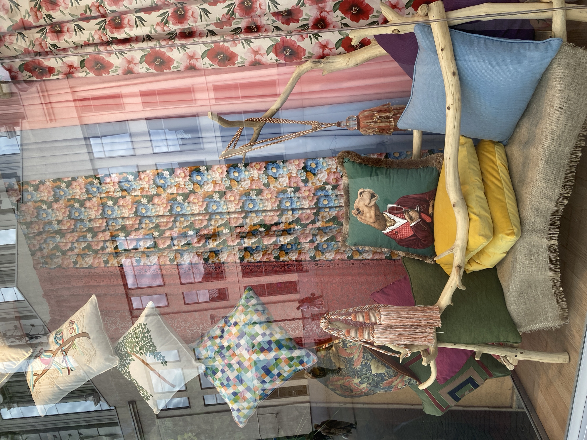 A window display contains cushions and fabric samples arranged on a wooden structure of branches. The cushions vary in color and design, including solid colors (blue, yellow, green, purple), floral patterns, a checkered pattern, and one with a drawing of a bird on a branch. Another has a drawing of a tree. One cushion has a print featuring a camel wearing a vest. The fabric samples are of various patterns and textures, including a floral print, a paisley print, and a linen-like weave. A tassel is hung across the display. The window reflects the exterior, showing buildings and other architectural details, suggesting an urban environment. Based on the buildings in the reflection, the location is Yekaterinburg, Russia.