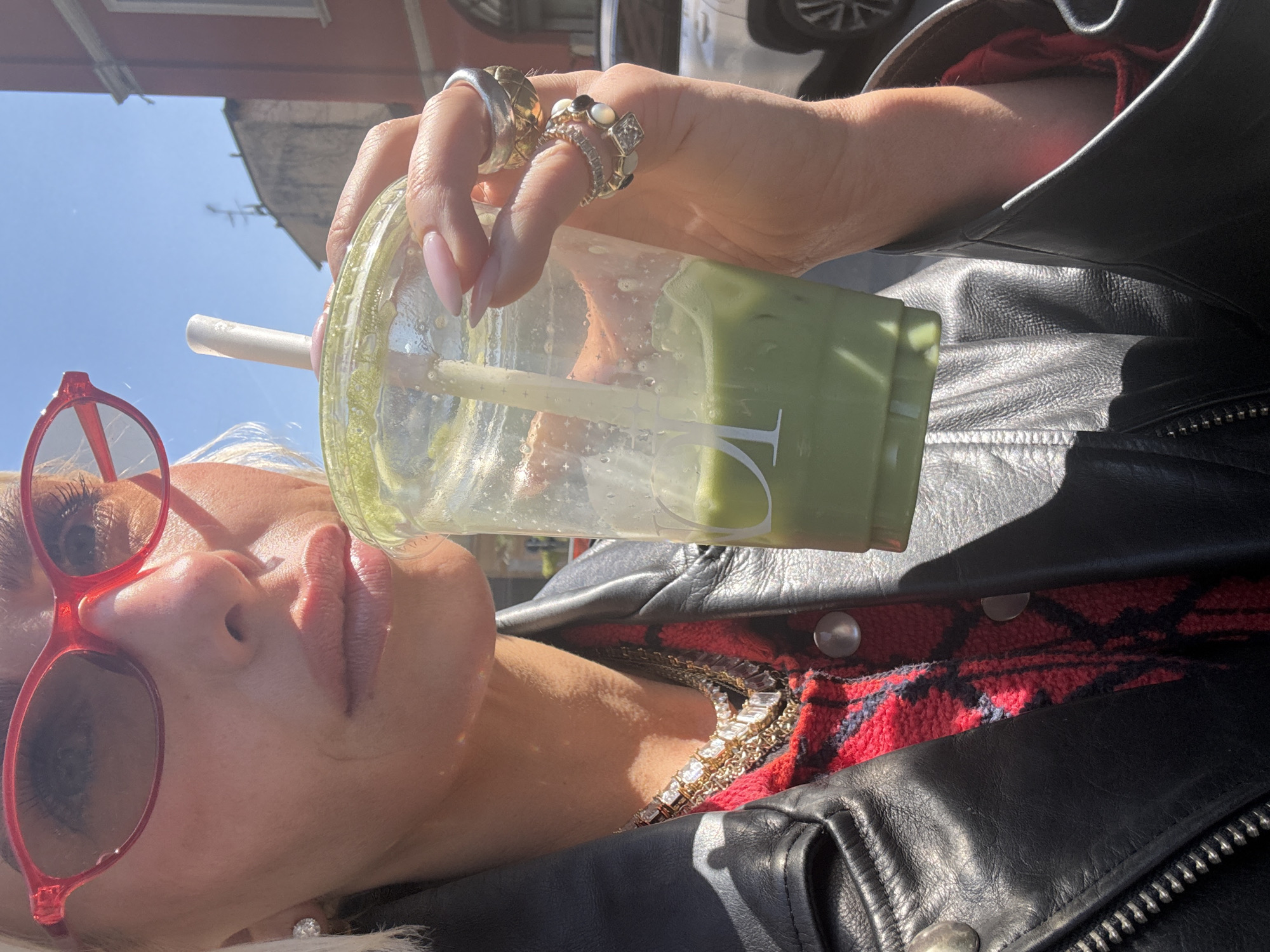 A woman is holding a cup of green liquid with a straw, possibly matcha latte. She has light skin, pale lipstick, and wears red sunglasses. She has several rings on her fingers and wears a black leather jacket over a red plaid shirt. A gold and diamond necklace is visible. The image appears to be taken outside on a sunny day. The location is Milan, Italy based on "PL" on the cup.