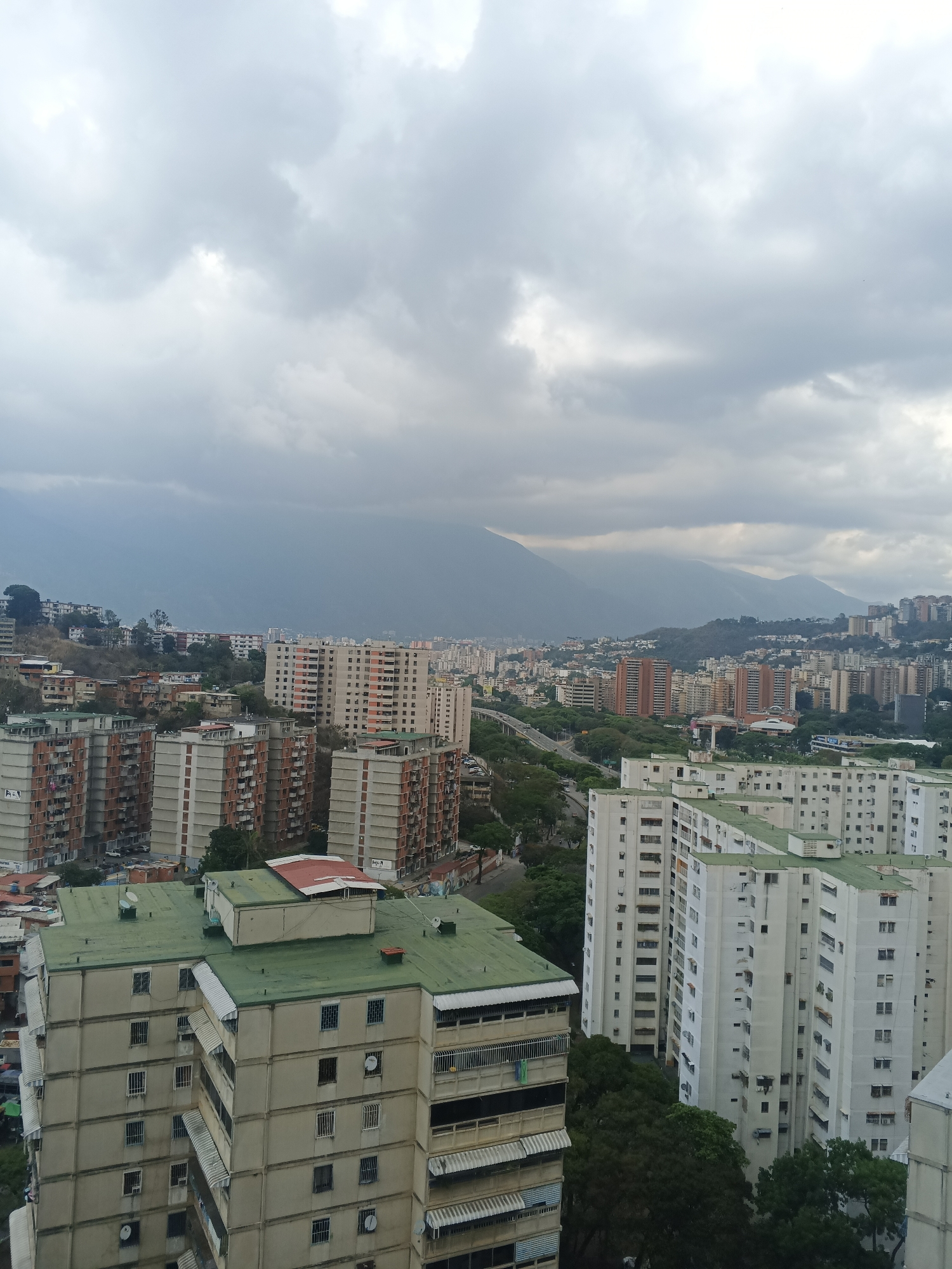 The image presents a wide, elevated view of a sprawling urban landscape in Caracas, Venezuela, under a heavily overcast sky. The scene is dominated by numerous multi-story residential buildings, extending from the immediate foreground to the distant horizon, with a backdrop of large mountains partially obscured by clouds and haze.
In the foreground and mid-ground, distinct clusters of apartment buildings are visible. On the left, several structures feature light-colored walls with prominent reddish-brown sections and flat green roofs. These buildings appear to be part of a large complex, with some showing balconies and security bars on windows. Faint text, possibly "A-1," is discernible on the lower-left side of one of these buildings. On the right side of the foreground, another large complex of light-colored, tall apartment buildings with green roofs is visible, displaying numerous windows, balconies, awnings, and satellite dishes. Green trees are scattered throughout the urban environment, breaking up the concrete landscape.
Further into the background, a busy cityscape unfolds with a mix of residential and commercial structures of varying heights and architectural styles. A road or highway can be seen winding through the mid-ground, connecting different parts of the city. Beyond the urban sprawl, majestic mountains rise, their upper slopes and peaks disappearing into the thick, gray cloud cover, suggesting either an impending rain or a typically cloudy day.
The time of day appears to be daytime, characterized by the diffused light from the overcast sky. There are no discernible people or specific activities taking place, offering a static perspective of the dense urban environment. The visible text "A-1" is noted on some of the buildings in the foreground.