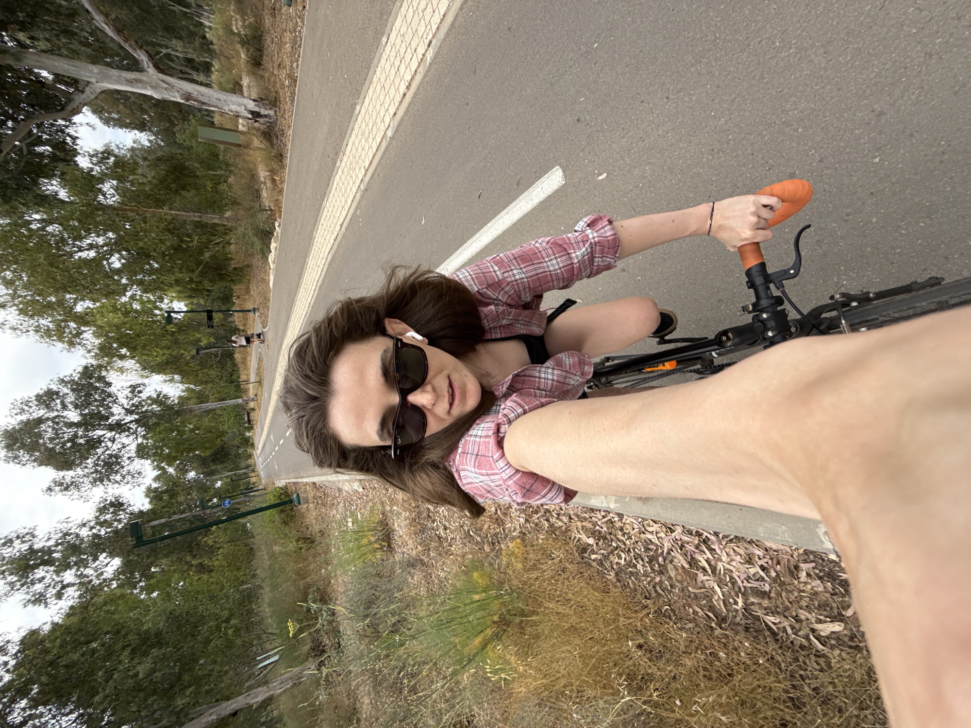 A woman wearing sunglasses and a pink plaid shirt is riding a bicycle on a paved path. She appears to be taking a selfie, with her arm extended towards the camera. The path has a textured lane marker, and the surrounding environment includes trees, vegetation, and what looks like dry leaves and grass. The location is consistent with a path in a park or green space, possibly in Tel-Aviv, Israel.