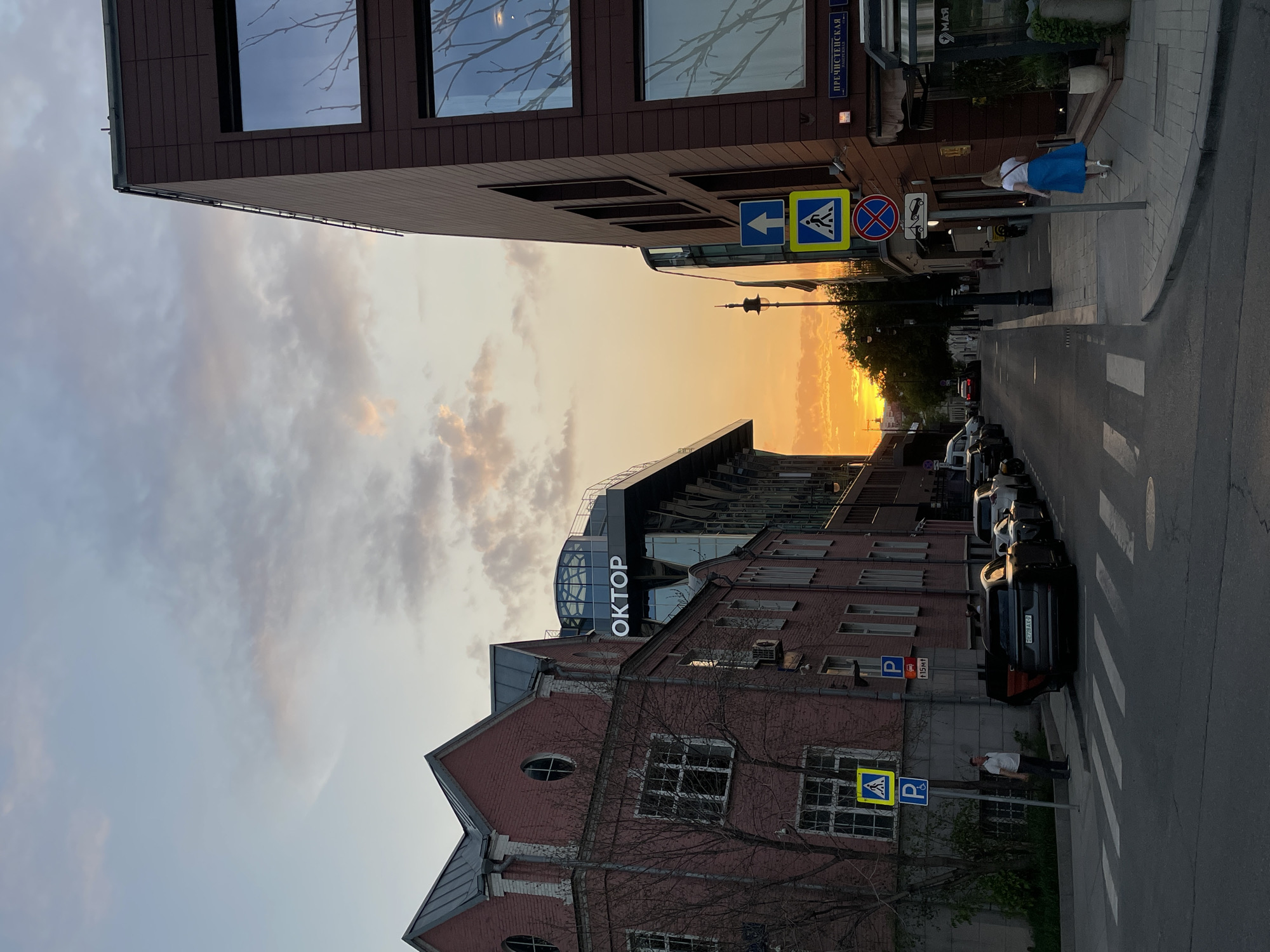 The image shows a street in Moscow, Russia at sunset. The street is lined with buildings, including one with the Cyrillic word "ОКТОР" visible. Cars are parked along the side of the road, and a crosswalk is visible. A person wearing a blue skirt is walking on the sidewalk near the upper building. There are street signs for pedestrians and street parking, and the building address reads "Пречистенская".