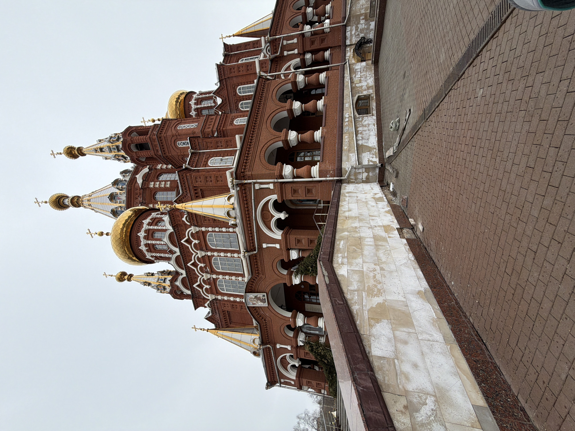 The image depicts St. Michael's Cathedral (Mikhailovsky Cathedral) in Izhevsk, Russia. The large, multi-tiered structure is constructed primarily of red brick, featuring extensive white decorative trim including arched windows, columns, and cornices. Multiple golden onion domes and pointed spires, each topped with a golden Orthodox cross, dominate the upper sections of the building against an overcast, light gray sky. An arched entrance with white columns is visible on the main facade, with an icon positioned above the central arch.
In the foreground, a reddish-brown brick paver walkway extends from the bottom of the frame, alongside a retaining wall composed of a dark reddish-brown granite base, topped by weathered light-colored stone panels, and capped with darker stone. A metal drainage grate runs across the paved surface. Near the wall, loose bricks and bags of material are visible on the pavement, suggesting minor ongoing work or debris. Bare deciduous trees are visible in the distant background to the left. No individuals are present in the scene. The lighting indicates an overcast day.