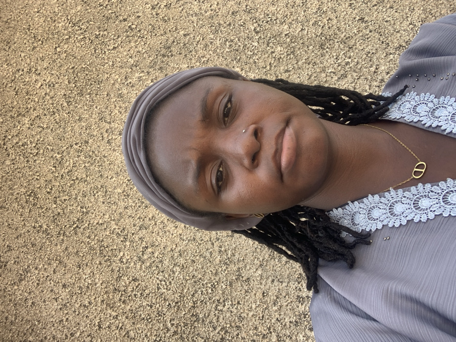 An adult female is depicted in a close-up, chest-up portrait shot against a light brown, textured wall in Bojude, Nigeria. She has dark skin, dark dreadlocks, and wears a grey headwrap covering her hair. Her attire consists of a grey garment featuring light blue or grey lace trim with small metallic stud embellishments along the neckline. A small silver stud pierces her left nostril, and a small gold-colored hoop earring is visible in her left earlobe. A gold-colored necklace with an interlocking oval or "CD" pendant rests on her chest. Her expression is neutral.