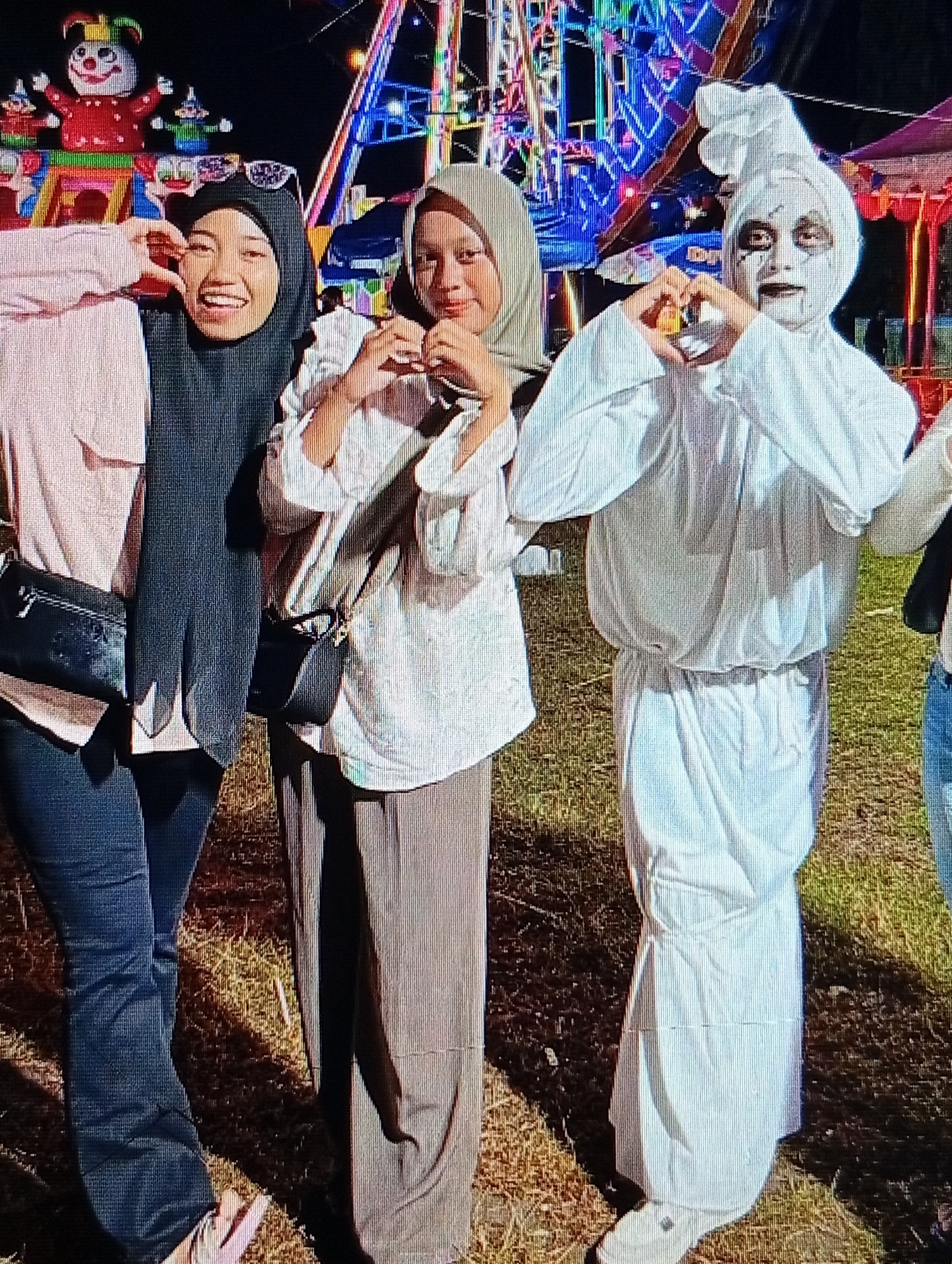 The image depicts three individuals at what appears to be an outdoor night event, possibly a carnival or fair, indicated by the colorful Ferris wheel-like structure in the background and amusement park figures. The person on the left is wearing a pink top, dark hijab, and dark pants, with a black crossbody bag. The person in the middle is wearing a light-colored hijab and a loose-fitting white top with dark pants. The person on the right is dressed in a white costume, resembling a ghost, with face paint that includes dark eye makeup and lips. All three individuals are making heart shapes with their hands, suggesting a friendly or celebratory interaction. The ground is covered with dry grass. The presence of the Ferris wheel and carnival elements suggests a public entertainment venue. It is not possible to determine the specific location in Lhokseumawe, Indonesia, from the provided image alone, as there are no identifiable landmarks or signs.