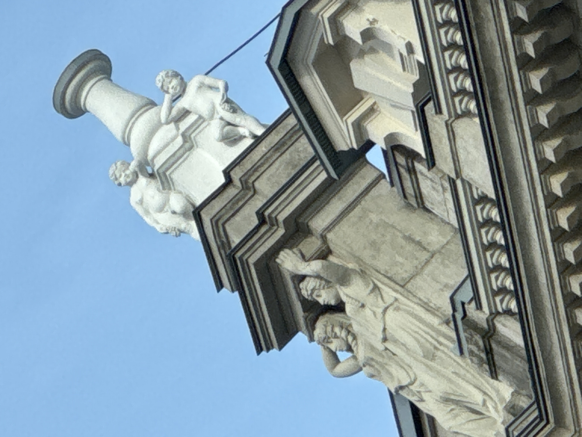 An upward-angled view captures ornate architectural details and classical statuary against a bright, clear blue sky. On the left, a large, white, decorative column-like structure features two cherubic or putto figures. One is lounging, resting its head on an arm, while another is positioned lower down. To the right and below, part of a building's facade is visible, adorned with intricate stone carvings. Two draped figures, possibly caryatids or atlantes, are depicted supporting an architectural projection with their hands and shoulders. These figures, sculpted in a classical style, show detailed drapery and form. The building itself exhibits elaborate cornices, moldings, and patterned friezes, suggesting a Baroque or Neoclassical architectural style. The material appears to be weathered light grey stone or stucco for the building, contrasting with the whiter statues. The sky is uniformly blue, indicating daytime and good weather. No text is visible in the image. The grand and detailed architecture is characteristic of historic buildings found in cities like Saint Petersburg, Russia.