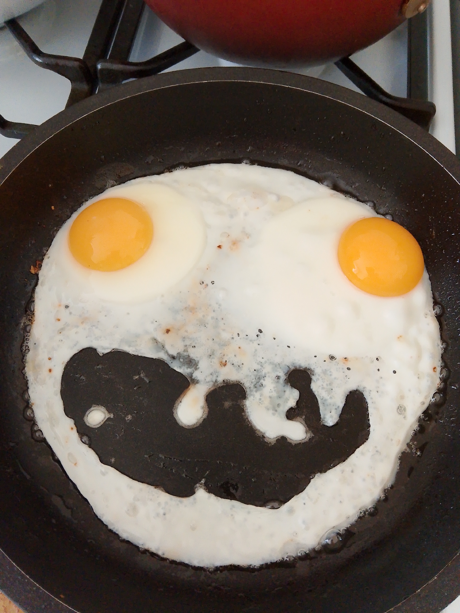 A top-down, eye-level shot shows two fried eggs in a dark non-stick frying pan, resembling a smiley face. The yolks are round and orange, positioned towards the top, resembling eyes. The cooked egg whites form a wide, amorphous shape that gives the impression of a mouth. The pan sits on a stovetop gas burner. Part of another red-brown colored pot is seen at the top of the frame. The environment suggests an indoor kitchen setting. Фоки, Russia is not detectable in the image.