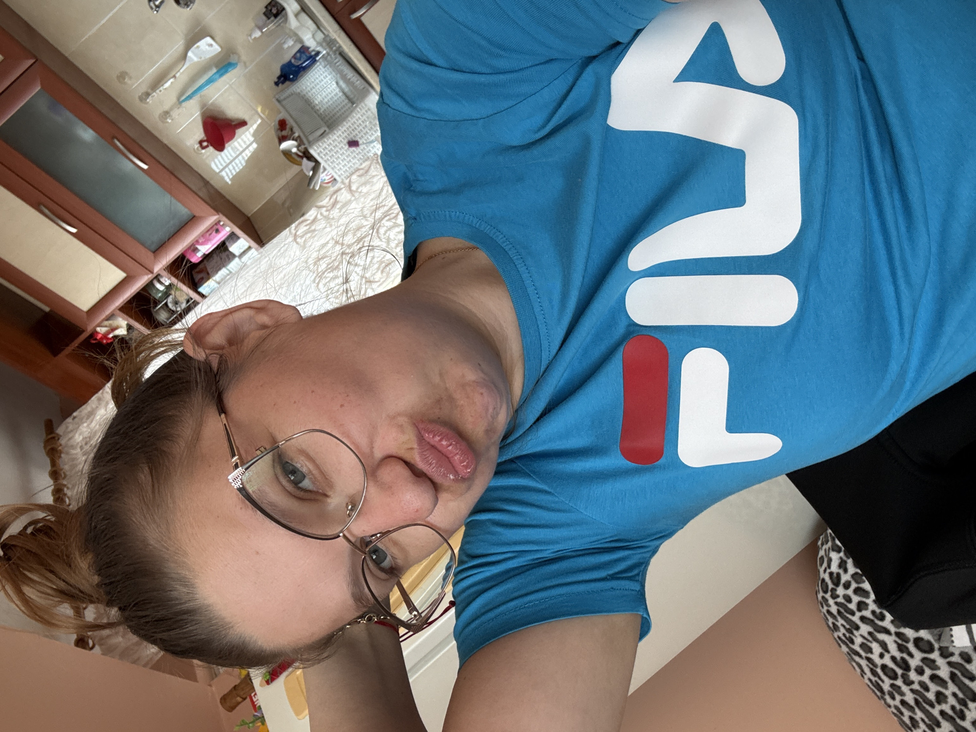 A woman wearing glasses and a blue Fila T-shirt poses for a selfie indoors. The background shows a room with a bed, some furniture, and what seems to be kitchen utensils and cabinets. The location is likely not in Sofia, Bulgaria, as there is no indication of it in the provided image.