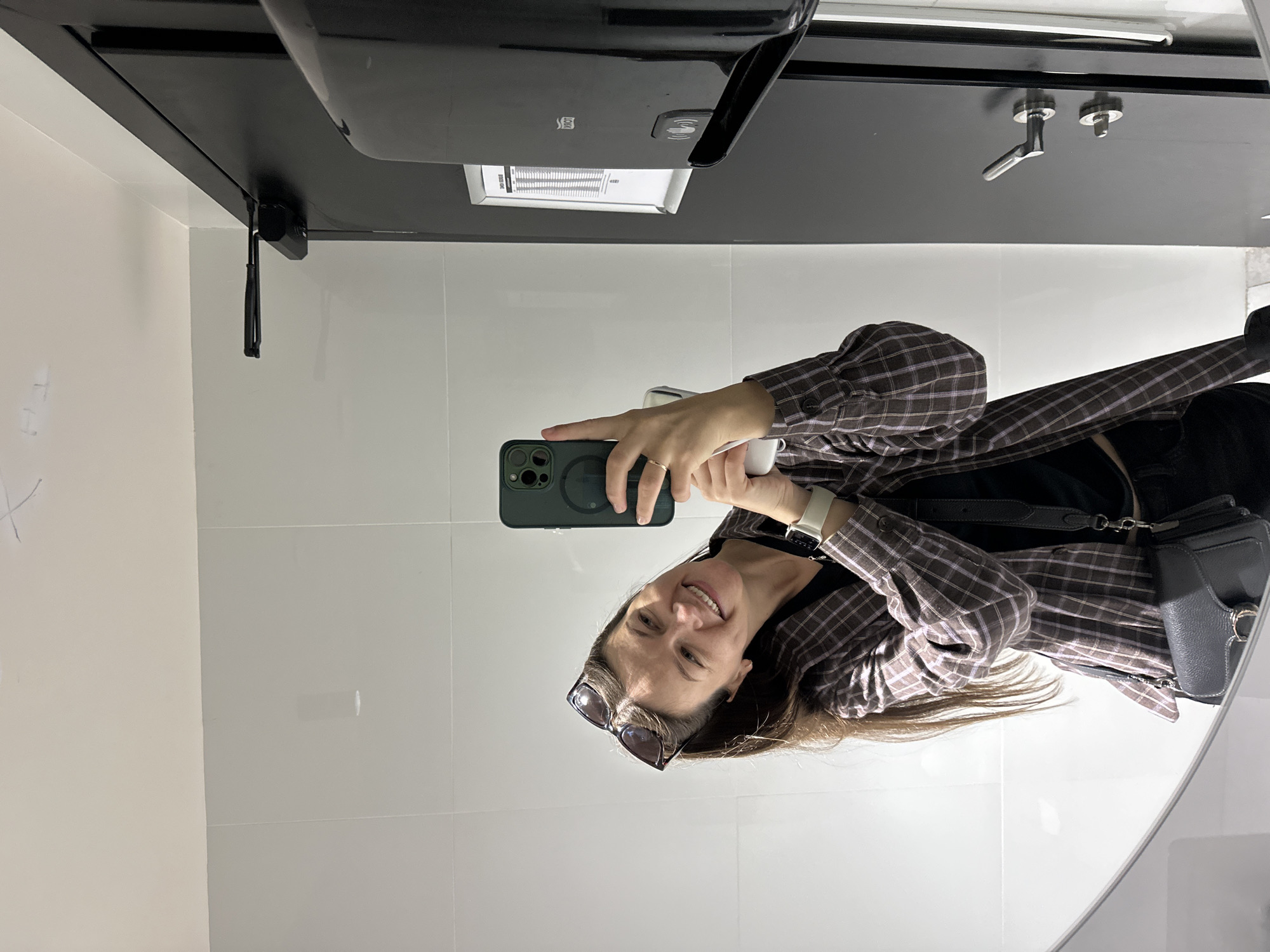 The image shows a woman taking a selfie in a bathroom. She is holding an iPhone in her left hand and appears to be smiling. She is wearing a plaid shirt, a black top, and sunglasses on her head. She is wearing a smartwatch on her left wrist. A dark gray crossbody purse is visible on her right hip. The bathroom has white tiled walls and a black countertop. A paper towel dispenser is attached to the wall above the countertop. The sink has a modern faucet. Based on the tiles and fixtures, this may be a public restroom. Although I cannot determine the exact location based on the provided image, the prompt indicates it is in the city of Budva, Montenegro.