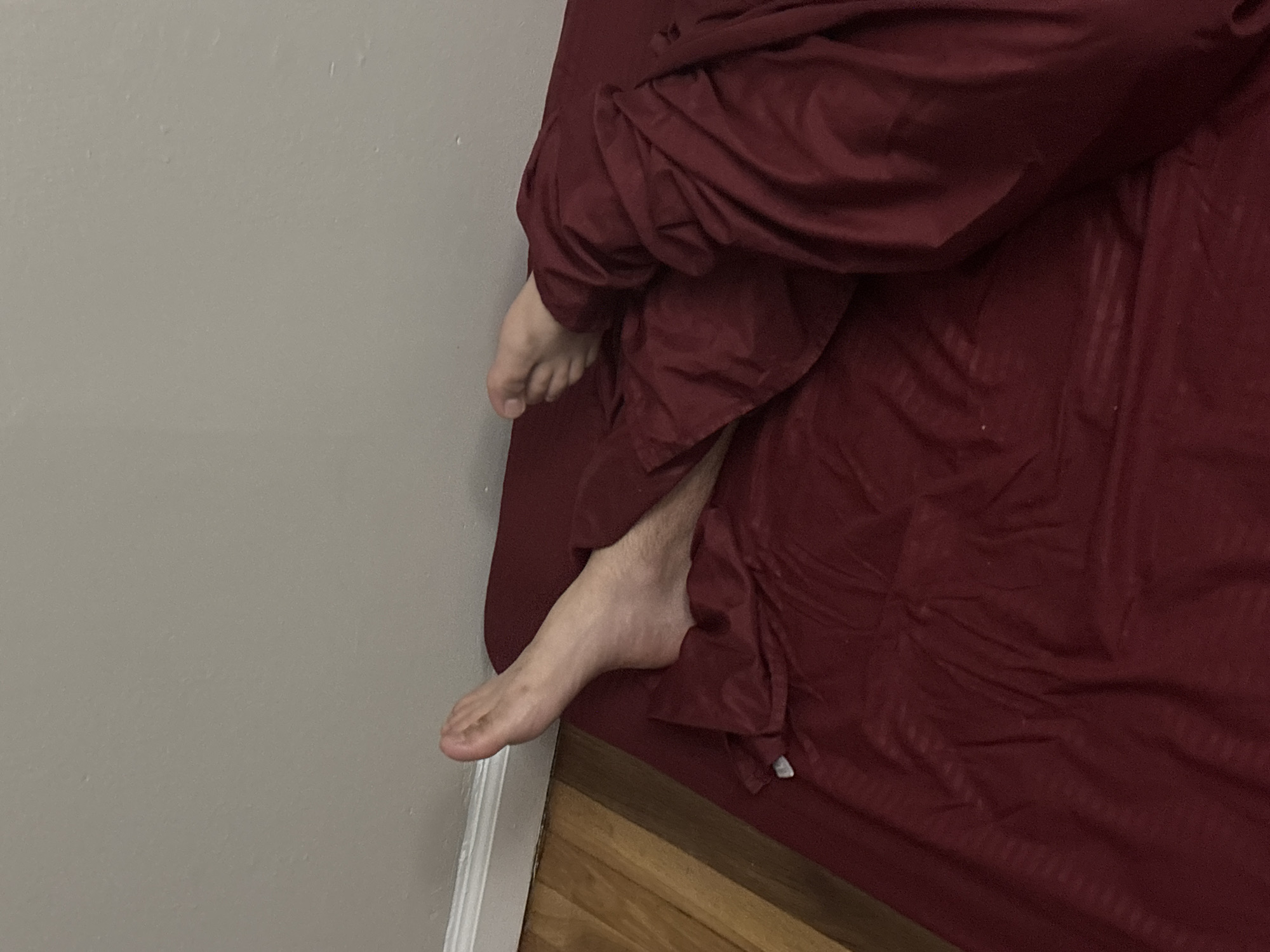The image captures an indoor scene, likely a bedroom, from an elevated perspective looking downwards. The primary subjects are the bare feet and lower legs of a person, partially covered by dark red or maroon bedding. One foot, with its toes and part of the top visible, emerges from the covers near the upper right of the frame. The other foot and a portion of the lower leg, displaying light skin and some sparse hair, extend further out from under the bedding towards the bottom left, resting close to a light gray wall.
The bedding itself is a solid dark red or maroon color, exhibiting a subtle vertical striped pattern and appearing rumpled, suggesting a person is either resting within it or has recently been in the bed. This bedding occupies the right side of the image. The left side is dominated by a plain, light gray or beige wall, which shows minor imperfections on its surface. At the bottom of the image, a section of wooden floor is visible, along with a white baseboard marking the transition between the wall and the floor.
The activity depicted is a person resting or sleeping, with their feet exposed from the covers, a common position for comfort. The scene suggests a private, relaxed, and domestic setting. There are no direct indications of the time of day, weather, or explicit emotions. No text is visible within the image. The context places this scene in Franklin Township, United States of America.