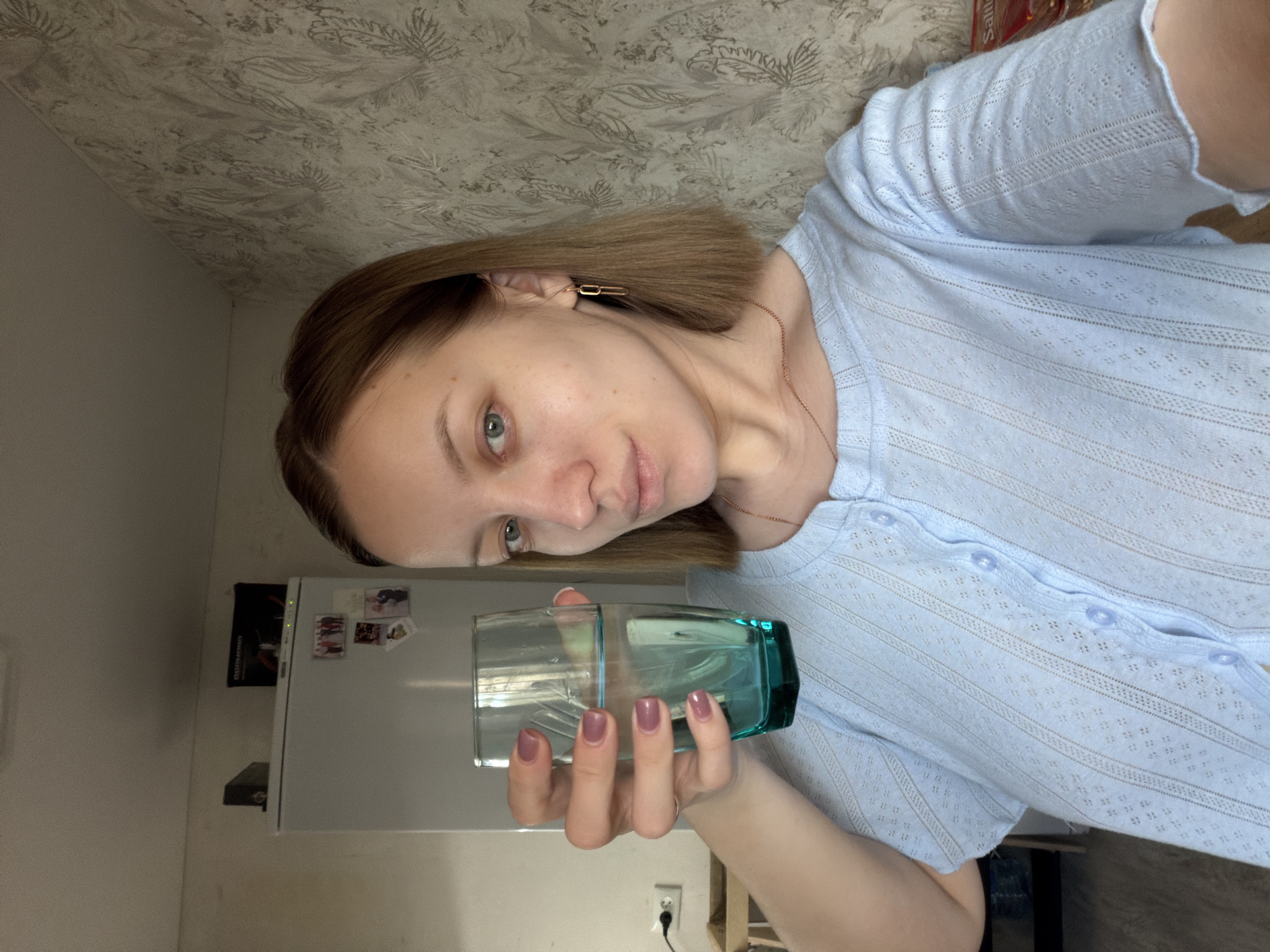 A young, fair-skinned woman is holding a glass of water in front of a light-colored wall. She has short, brown hair and is wearing a light blue, button-up shirt. The glass of water is partially clear and partially teal. In the background, there is a white refrigerator with some magnets and other items attached. A wall with a patterned wallpaper is visible above the woman. It is not possible to determine the precise location in the city of Reshetnikovo, Russia from the given image.