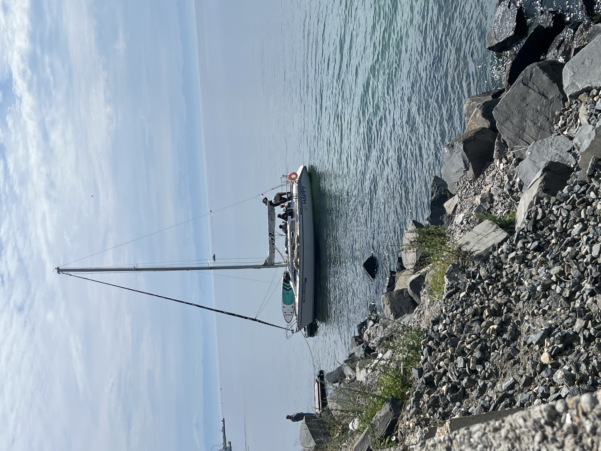 A sailboat, labeled "HAPPY," is moored near a rocky shore in Listvyanka, Russia. The boat has several people on deck, appearing to be engaged in leisure activity. A person stands on a wooden dock, seemingly observing the boat. The scene is set against a backdrop of calm water and a hazy sky, with distant mountains or hills visible on the horizon. The foreground is dominated by a rugged, rocky embankment.