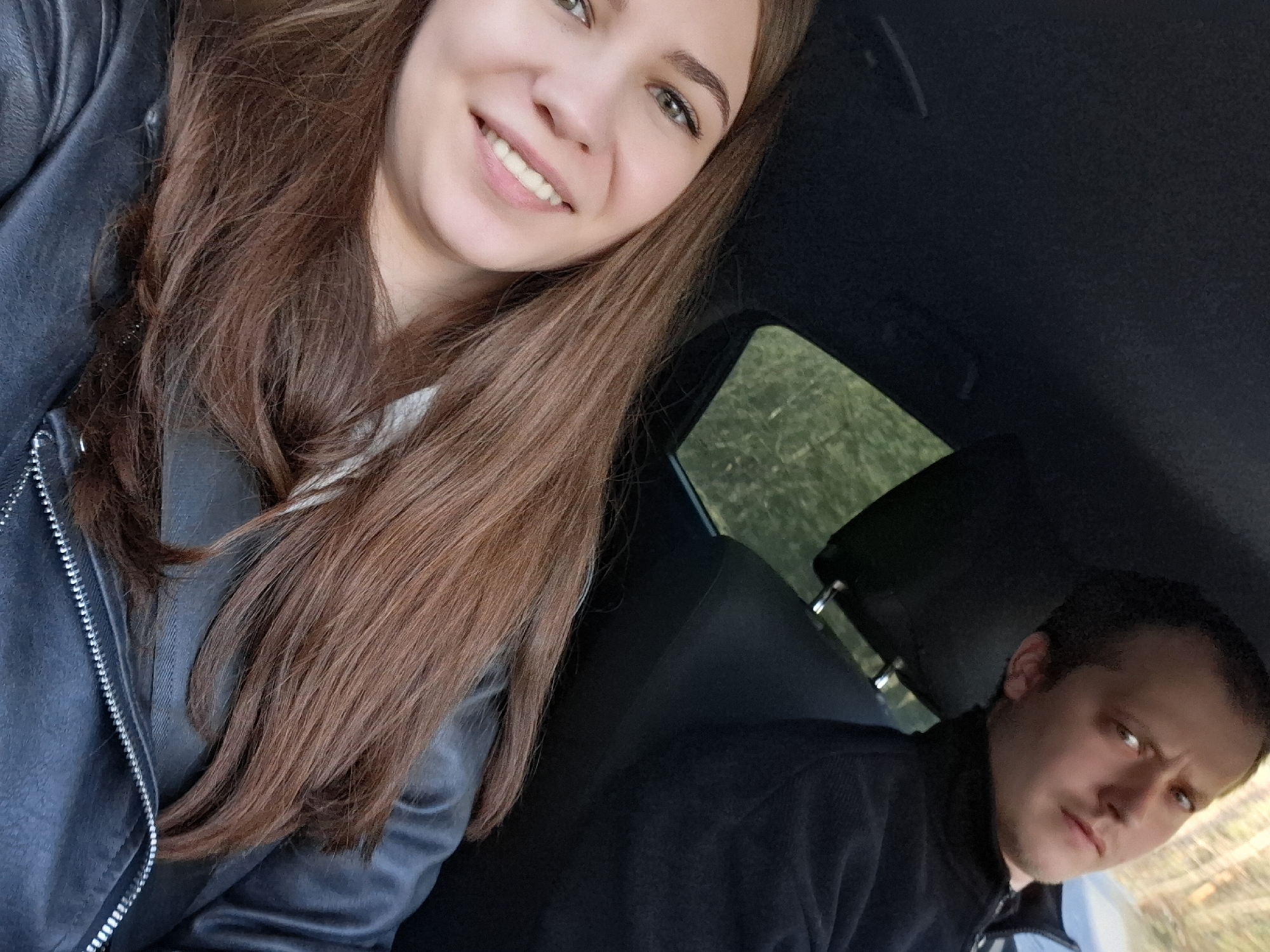 The image shows two individuals inside a vehicle, likely a car, with a green, tree-filled landscape visible through the back window. A young woman with long, brown hair, wearing a black leather jacket, is in the foreground taking the picture, and smiling at the camera. In the back is a man looking at the camera with a slight frown. The location is presumed to be Chaikovsky, Russia.