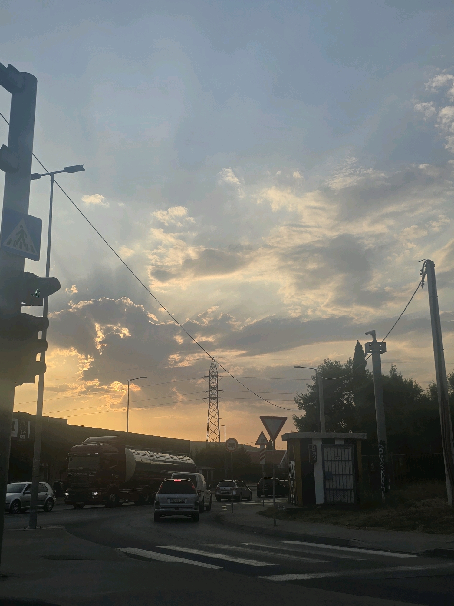 The image depicts a street scene in Podgorica, Montenegro, during what appears to be sunset or sunrise. The sky is filled with dramatic clouds, illuminated by the warm hues of the setting or rising sun. In the foreground, a road intersection is visible with a pedestrian crossing. Several vehicles are present: a large red tanker truck, a silver car, and a silver SUV with its brake lights illuminated. A traffic light is mounted on a pole on the left side of the image, alongside a pedestrian crossing sign. In the background, there are buildings, trees, and utility poles, including a prominent electricity pylon. The overall atmosphere is one of urban movement under a striking sky.