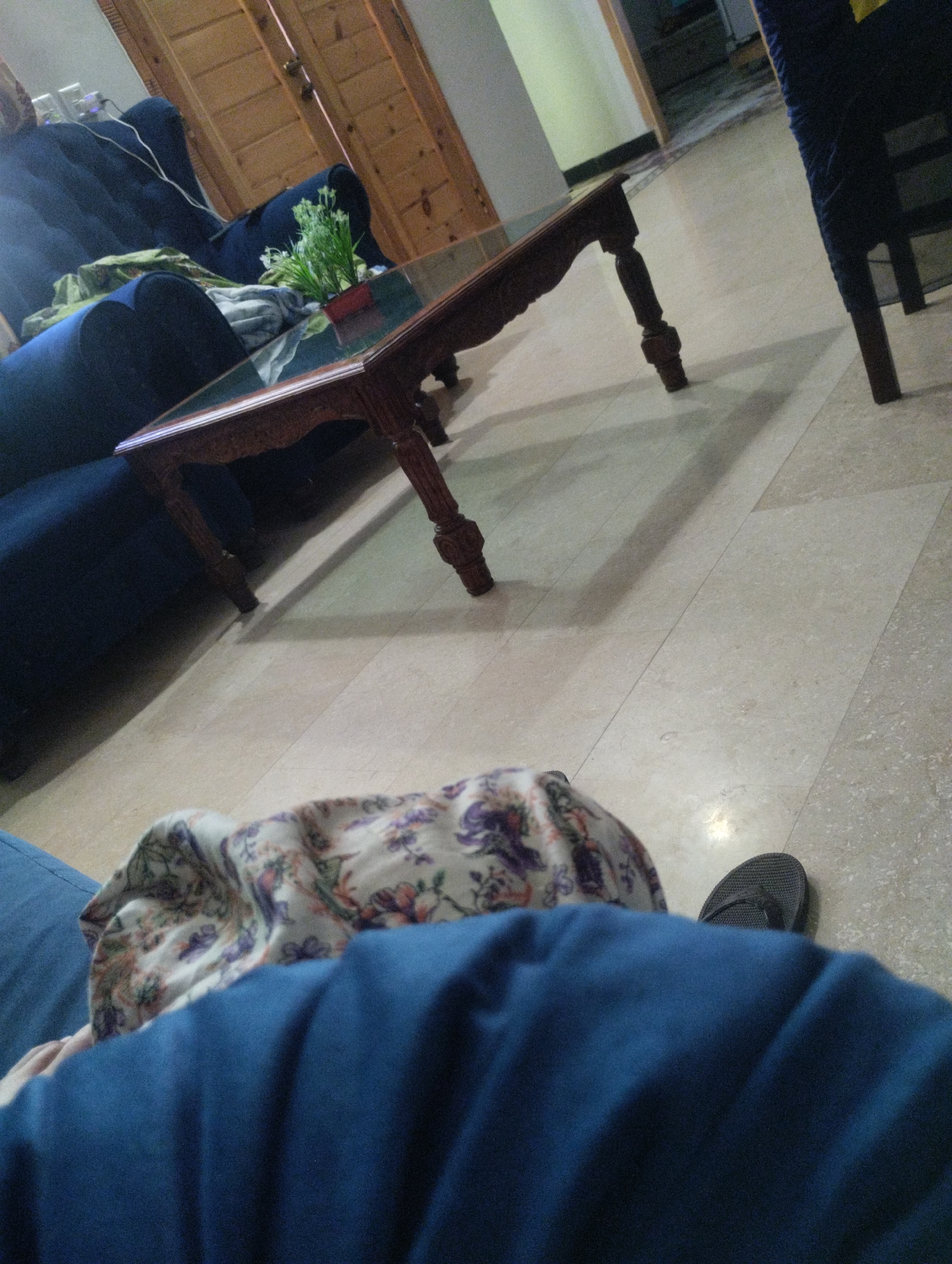The image depicts a dimly lit indoor scene. In the foreground, a person's legs are visible, covered by blue fabric, with a patterned cloth draped over them. A single black flip-flop is visible on the floor to the right of the person's legs. In the middle ground, a dark wooden coffee table with intricate carvings sits on a light-colored tiled floor. A glass top is visible on the table. Behind the coffee table, a portion of a dark blue sofa is visible, with what appears to be some fabric draped over it. To the left of the sofa, there are what seem to be electrical outlets on a wall. Further back, a wooden door with vertical panels is visible, and a doorway leads to another room where a portion of a dark chair is seen. The environment appears to be a living room or lounge area.