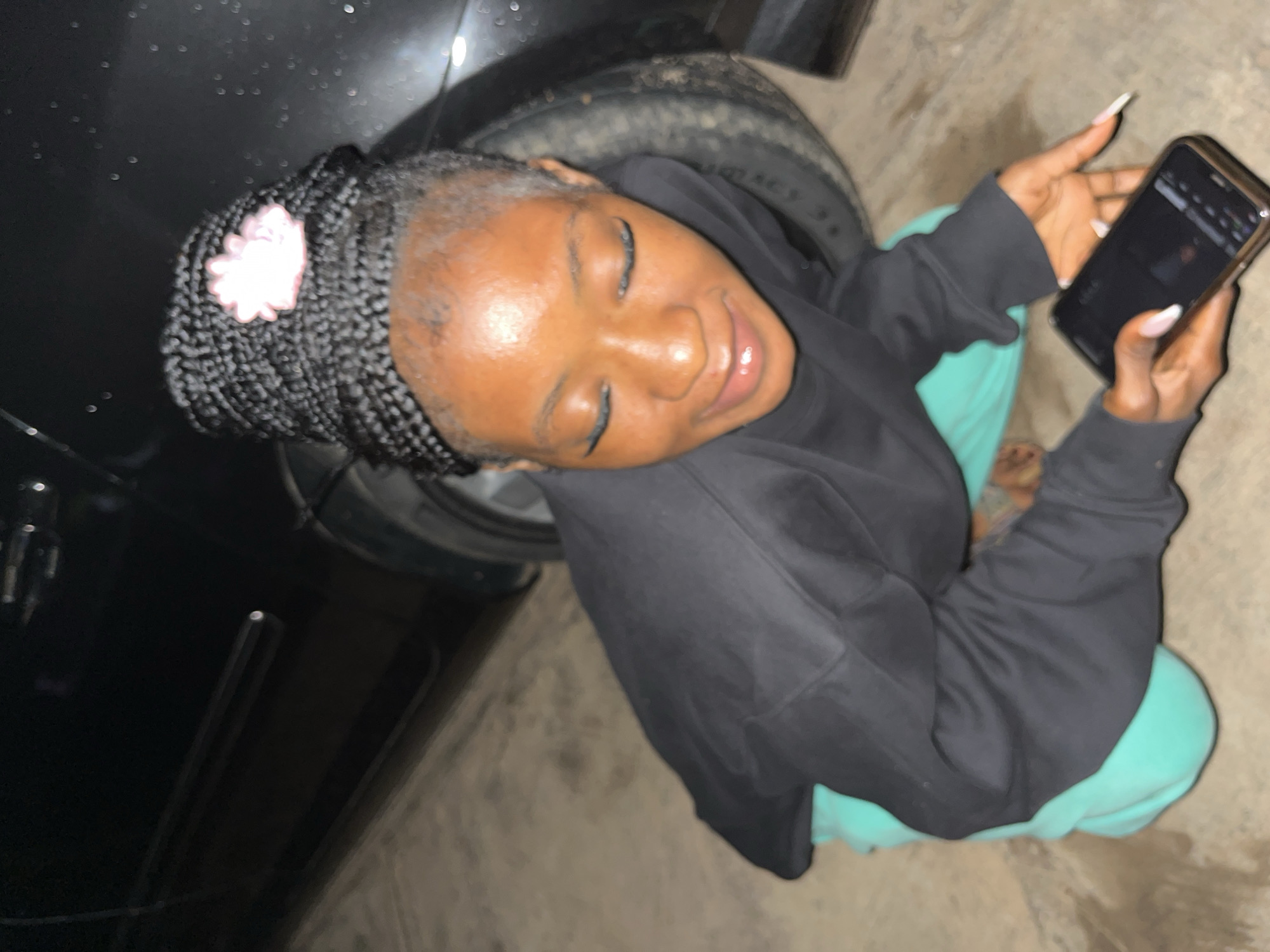 A young woman with braided hair adorned with a pink flower is crouching next to the rear tire of a dark-colored vehicle, possibly a car, on a paved surface that appears to be a street or parking lot in Jos, Nigeria. She is wearing a black sweatshirt and teal-colored sweatpants and is holding a smartphone in her right hand, appearing to interact with it. The lighting suggests it might be evening or nighttime, and the ground shows some signs of dampness.