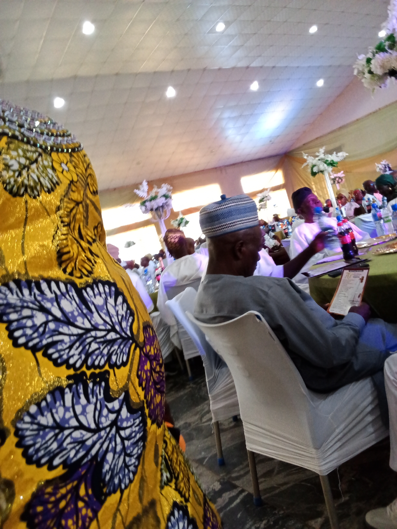 The image depicts an indoor gathering, likely a celebration or event, with an emphasis on the seating arrangements and attendees. In the foreground, a person's shoulder and arm are visible, adorned in a vibrant yellow fabric with a striking purple and white leaf pattern. The fabric appears to be richly textured, possibly embellished with gold threads.
The middle ground of the image focuses on a seated man wearing a blue and white patterned cap and a grey tunic. He is holding a document and appears to be engaged with it, possibly reading or reviewing information. Another individual in the background, also in white attire and a cap, is reaching for a bottle of water. Several other guests are visible, seated at tables covered with green cloths. Bottles of water and other beverages are distributed across the tables, suggesting a meal or reception is in progress.
The environment is characterized by a light-colored, tiled ceiling with recessed lighting fixtures. The walls are draped with light-colored fabric, possibly for decoration, creating a soft ambiance. Floral arrangements, composed of white flowers, are placed strategically as centerpieces on some tables and suspended from above, adding to the festive atmosphere. The floor appears to be tiled with a dark, possibly polished, surface. The overall impression is of a well-organized event in a spacious indoor venue, with a focus on social interaction and dining.