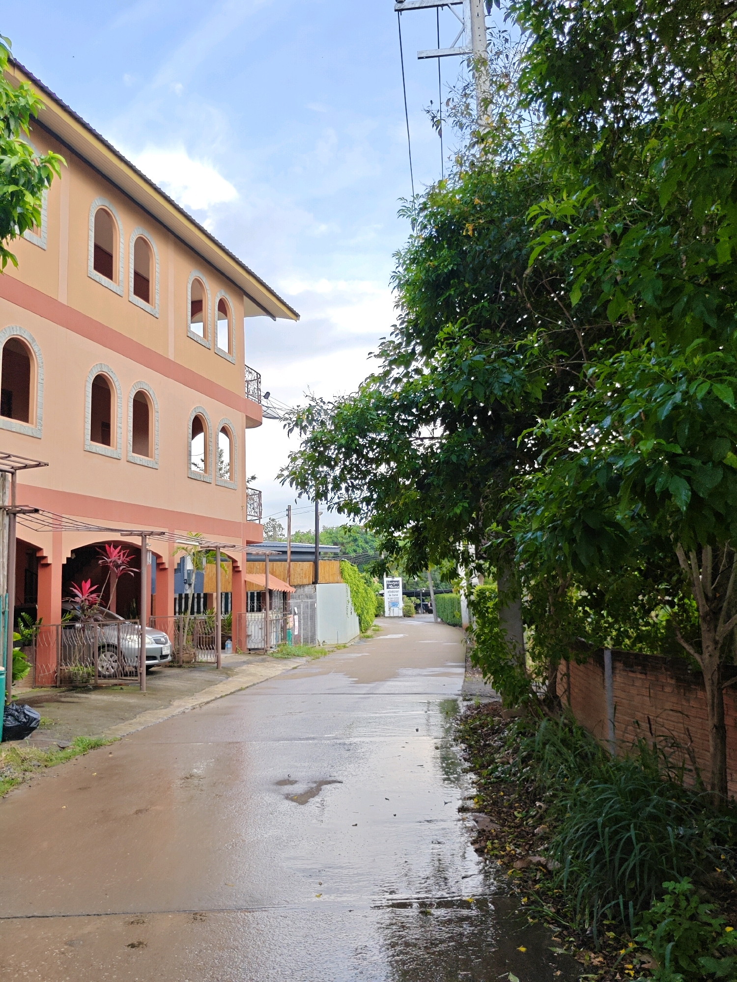 This image captures a street scene in Mu Si, Thailand, following a period of rain. The road surface is wet and reflective, with visible puddles. On the left side of the street, a multi-story building with peach-colored walls and arched windows is prominent. The lower level of this building features a covered parking area with a silver car parked beneath it. A fence separates the property from the street. To the right, a dense line of green trees and vegetation runs along the side of the road, with a brick wall visible behind some of the foliage. A utility pole with wires is also present in the background, contributing to the urban or semi-urban setting. The sky above is partly cloudy, with a light blue hue. The overall impression is of a residential or commercial street that has recently experienced wet weather.