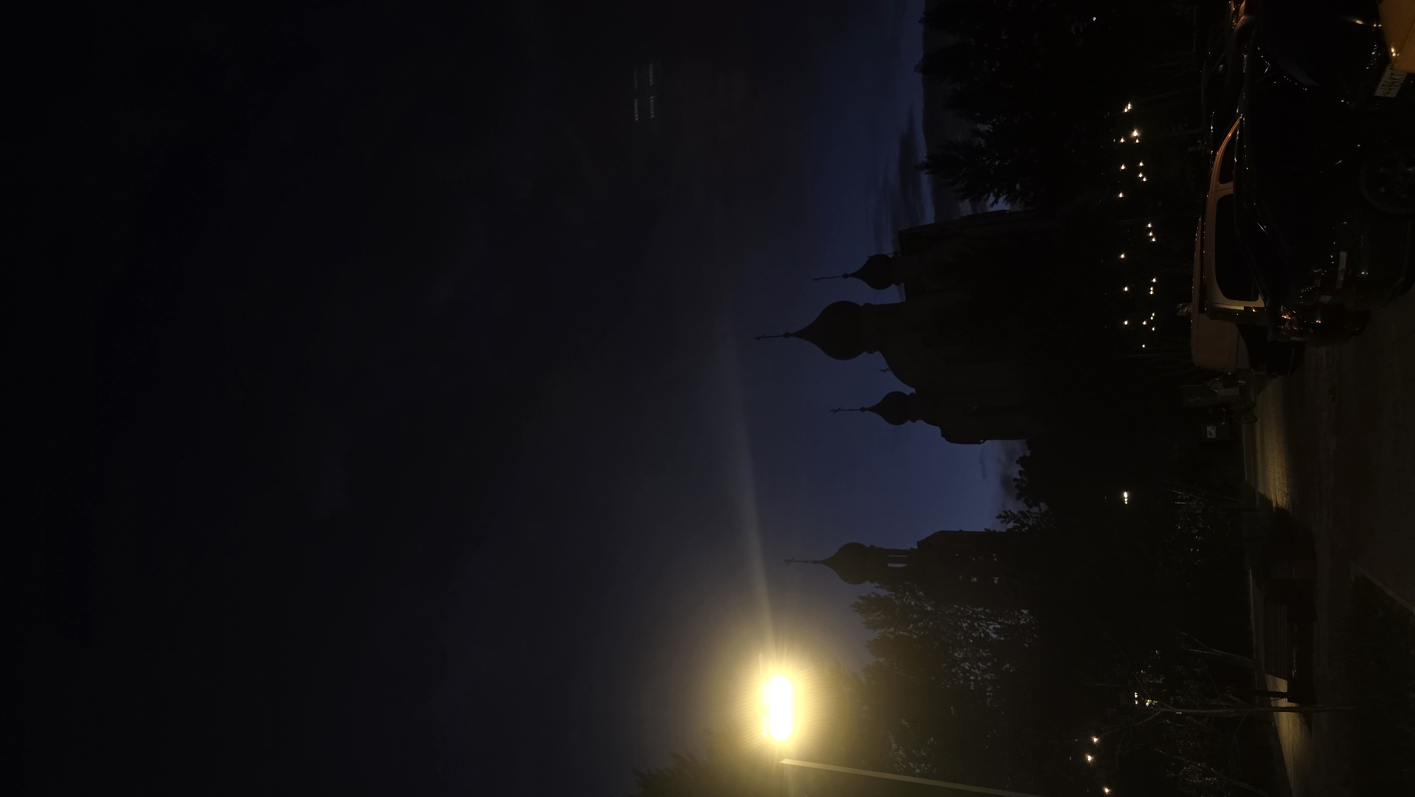 The image depicts an outdoor scene at dusk or night in an urban environment, with the sky transitioning from dark blue to black. In the background, the silhouette of a church with multiple domes and crosses is visible against the dim sky. A brightly lit street lamp is positioned on the left side of the frame, casting a strong yellow glow and creating lens flare. Silhouetted trees frame the church and the street lamp. In the foreground, several parked cars are visible, with their shapes and details obscured by the low light conditions. The ground appears to be paved, suggesting a parking lot or street area. The overall atmosphere is dark and somewhat eerie, with artificial light sources providing the main illumination. The location is described as Unknown, Unknown.