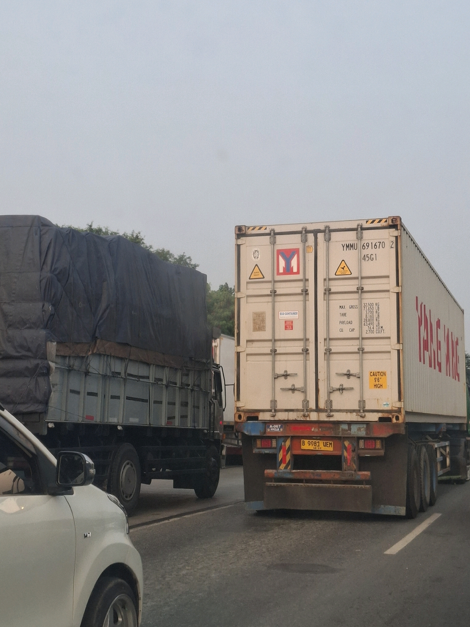 The image shows a street scene in Panunggangan Barat, Indonesia. In the foreground, the left side of a white car is visible, with its side mirror and part of its wheel arch. Behind the car, a large truck with a black tarp covering its cargo is traveling on the road. To the right of this truck, a white shipping container is mounted on a trailer, also in motion. The shipping container has various markings, including "YMMU 691670", "45G1", "MAX. GROSS 32,500 KG", and "CAUTION 9'6" HIGH". The license plate of the trailer is partially visible as "B 9981 UEM". Trees are visible in the background. The sky is overcast and a light haze is present. The overall impression is of traffic on a road, likely during the daytime.