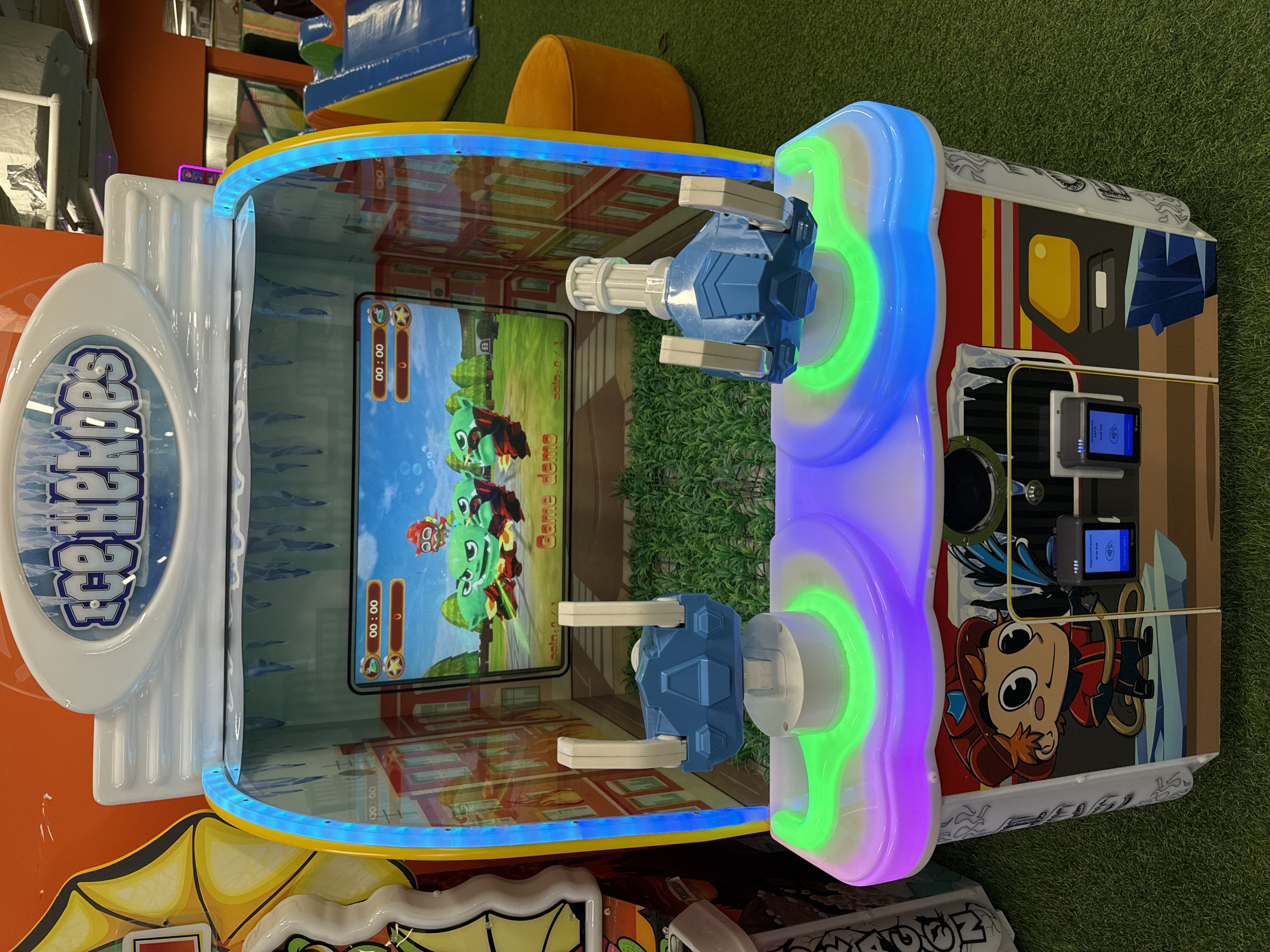 A close-up shot displays a "Fire Heroes" arcade game, situated on a green surface. The game features a screen displaying an animated scene and interactive turret-like controls. The exterior of the game is decorated with colorful graphics and illuminated with LED lighting. The game is indoors and located in the city of Khimki, Russia.
