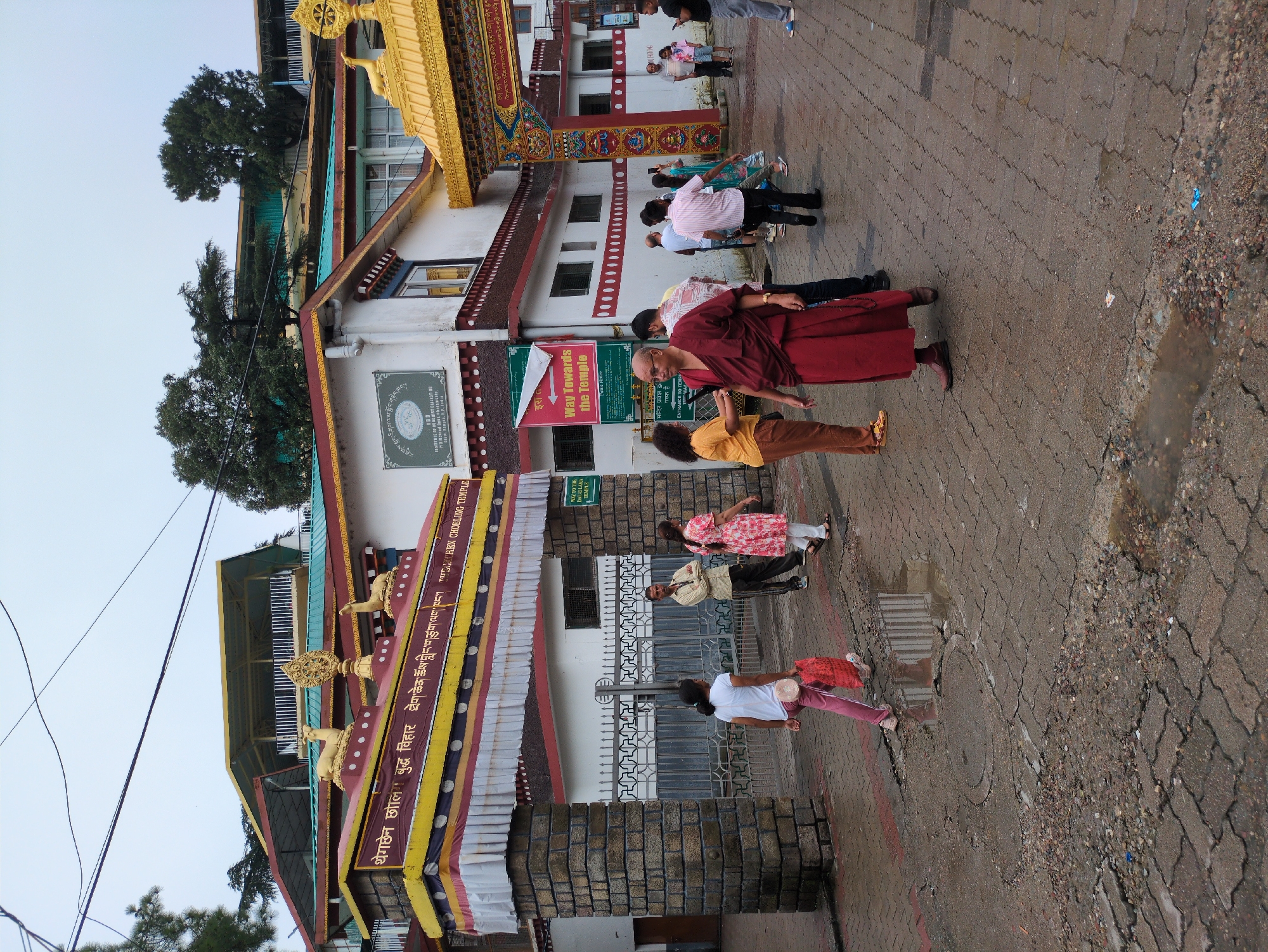 The image depicts the entrance to the Tenzing Memorial Museum in Dharamshala, India. The museum is part of the Namgyal Monastery, the Tibetan government-in-exile's monastic complex. In the foreground, a monk wearing a maroon robe walks towards the viewer, holding prayer beads. Several other people, including tourists and locals, are visible near the entrance, some taking photos and others observing the surroundings. The entrance itself is adorned with Tibetan Buddhist motifs, including golden deer statues and prayer flags. The architecture suggests a blend of traditional Tibetan and Indian styles. The ground is wet, indicating recent rainfall, and the sky is overcast. The overall scene conveys a sense of religious and cultural significance, with a peaceful atmosphere despite the presence of visitors.