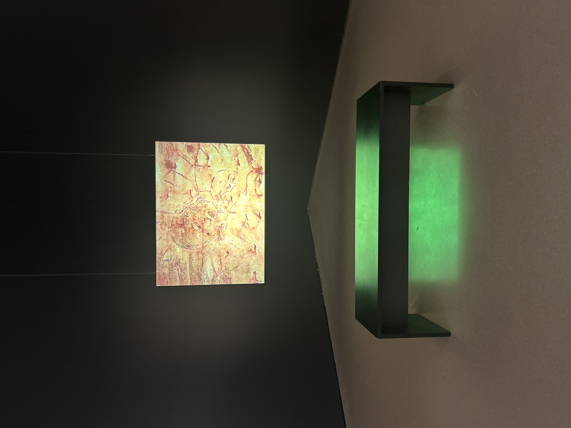 An indoor scene showcases an artistic or historical exhibit, likely within a gallery or museum. On the left, a rectangular artwork is prominently displayed against a dark, possibly black, wall. This piece, illuminated with a warm yellowish glow, features intricate patterns and textures in shades of yellow, orange, and reddish-brown, reminiscent of ancient art or highly textured abstract work. A thin, horizontal wire is visible, suggesting the artwork is suspended.
To the right, a lighter, beige-colored wall meets the dark wall at an acute angle, creating a dynamic architectural line. Mounted on this lighter wall is a dark, rectangular light fixture, possibly a sconce or shelf-like unit. This fixture emits a vivid green light, casting a distinct glow onto the wall surface behind and around it, creating a strong color contrast with the warm tones of the artwork. The overall lighting is dim, emphasizing the illuminated elements of the display. No people are visible, and there is no discernible text in the image. The arrangement suggests a curated display designed to evoke a contemplative atmosphere.