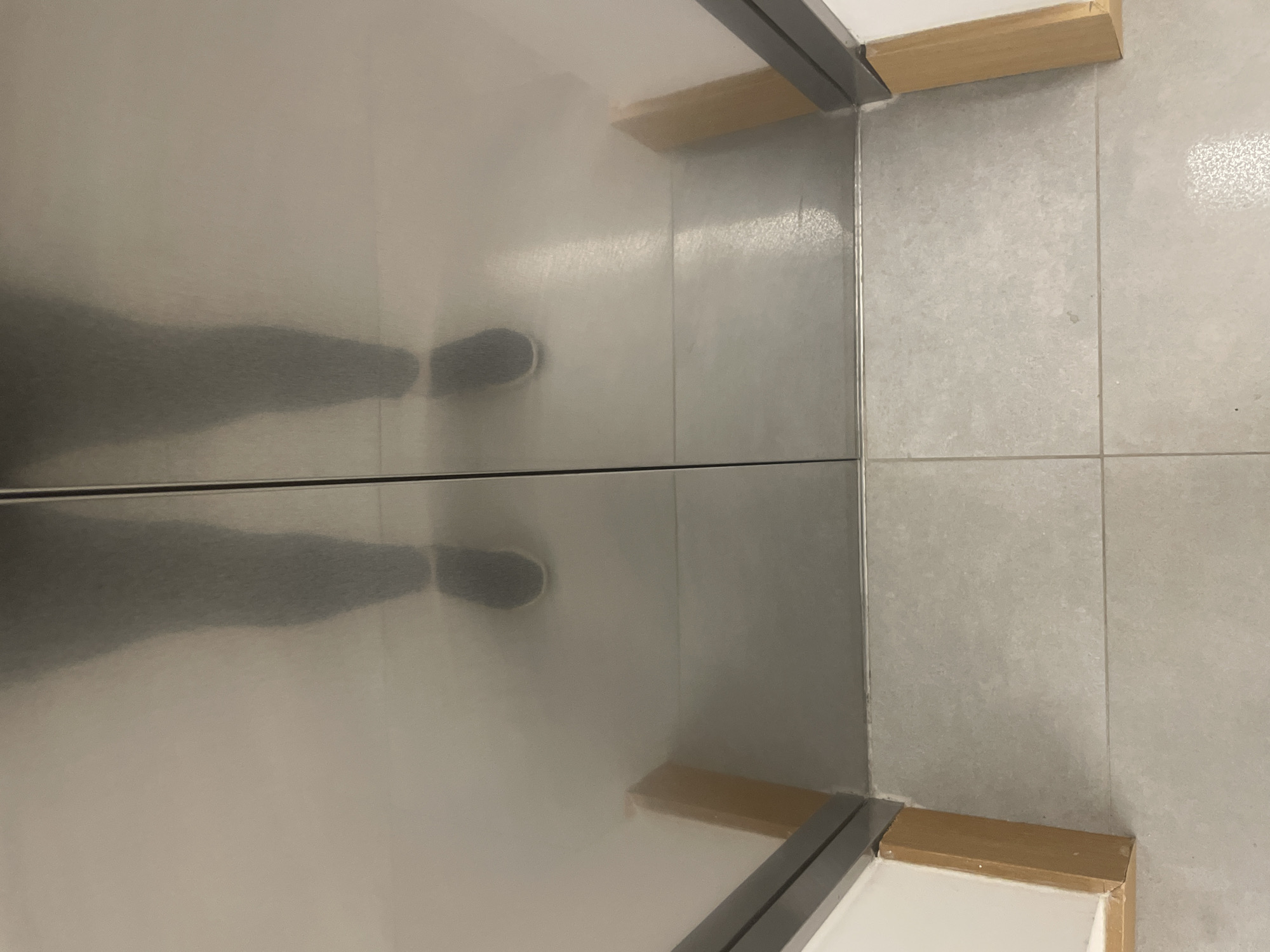 The image shows a reflection of a person's legs and feet in a mirrored surface. The person is wearing dark shoes and the reflection suggests they are standing on a tiled floor. The mirrored surface appears to be a wall or a partition, with a wooden support at the bottom. To the right of the mirrored surface, a wall covered with light-colored tiles is visible. The scene is indoors, likely in a modern setting such as a bathroom, changing room, or a shop. There are no discernible time of day or weather cues. The overall impression is neutral, with no strong emotional indicators. No text is visible in the image. The location context of Yunus Emre, Turkey, provides a geographical setting for this indoor scene.
