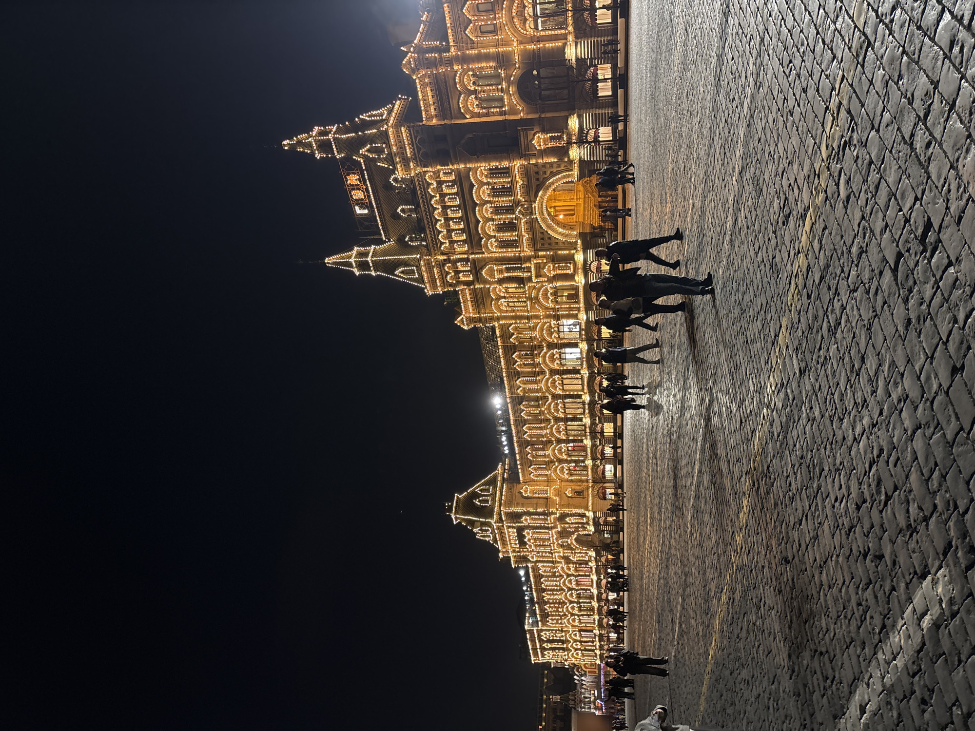 The image shows the GUM department store on Red Square in Moscow, Russia, at night. The building is illuminated with many lights. People are walking on the cobblestone square in front of the store. The sky is dark.