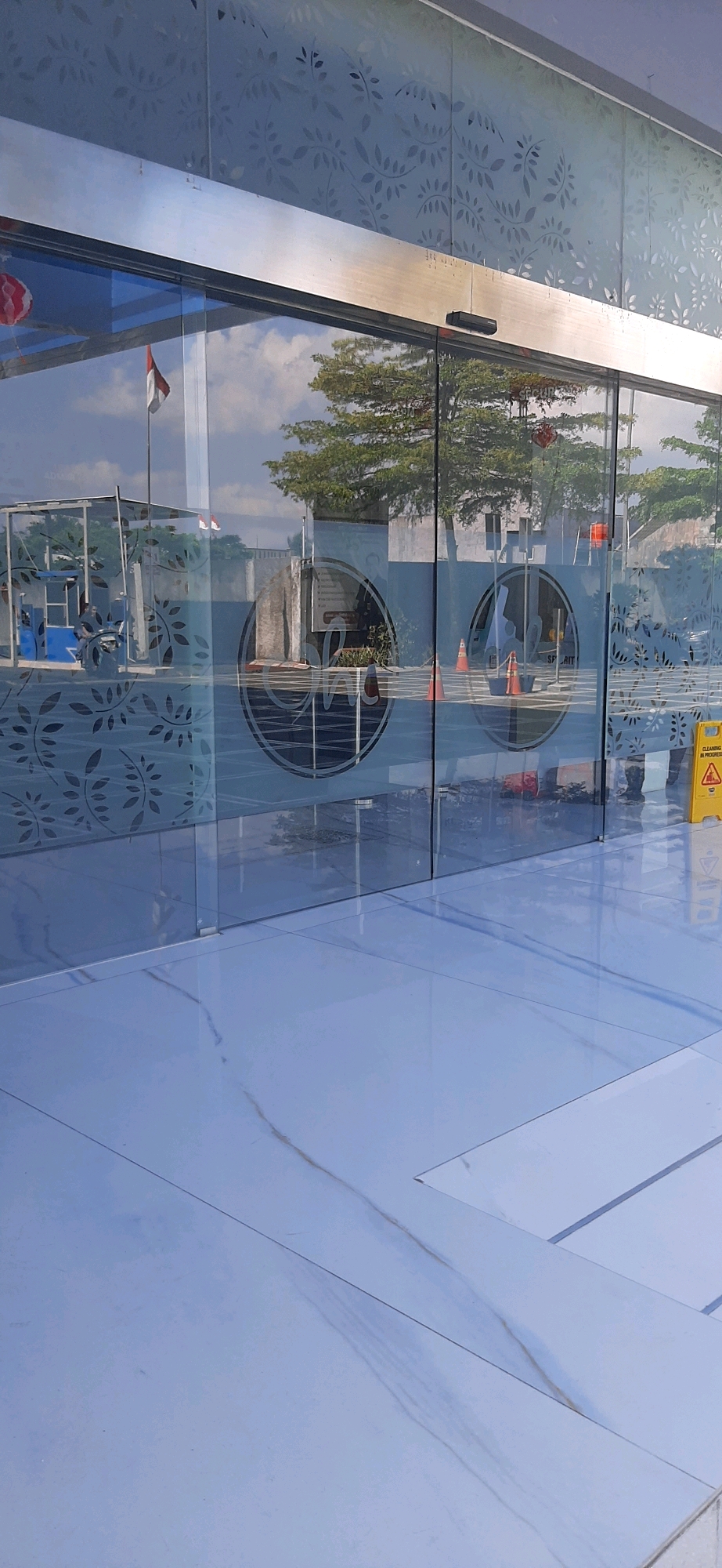 The image depicts the entrance of a building with automatic sliding glass doors. The doors are decorated with a subtle leaf pattern. Inside the building, reflections show a tree, a flag, and what appears to be part of a modern structure. On the floor outside the entrance, large, light-colored tiles with a faint veining pattern are visible. To the right, a yellow caution sign indicating "CLEANING IN PROGRESS" is placed on the floor. The overall scene suggests a commercial or public building entrance. The location is identified as the building of PT. Bank Central Asia Tbk. in Tasikmalaya, Indonesia.