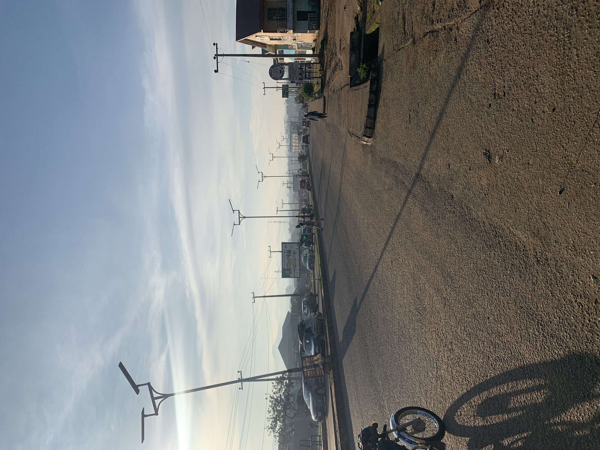 This is a forward-facing, eye-level, medium shot of a street in Rafin Par, Nigeria. The paved road is wide and lined with sidewalks on both sides. The street is populated with several cars and motorcycles, suggesting it is a busy thoroughfare. A few pedestrians are also visible, walking on the sidewalks or crossing the road. The scene is bathed in the soft, warm light of what appears to be early morning or late afternoon, casting long shadows across the road. The sky above is partly cloudy, with streaks of white clouds against a pale blue backdrop. In the background, buildings of various sizes and architectural styles line the street, some appearing to be shops or residential homes. Further in the distance, hills or mountains are visible, shrouded in a light haze. The overall impression is that of an active urban environment in Nigeria, captured during a peaceful time of day.