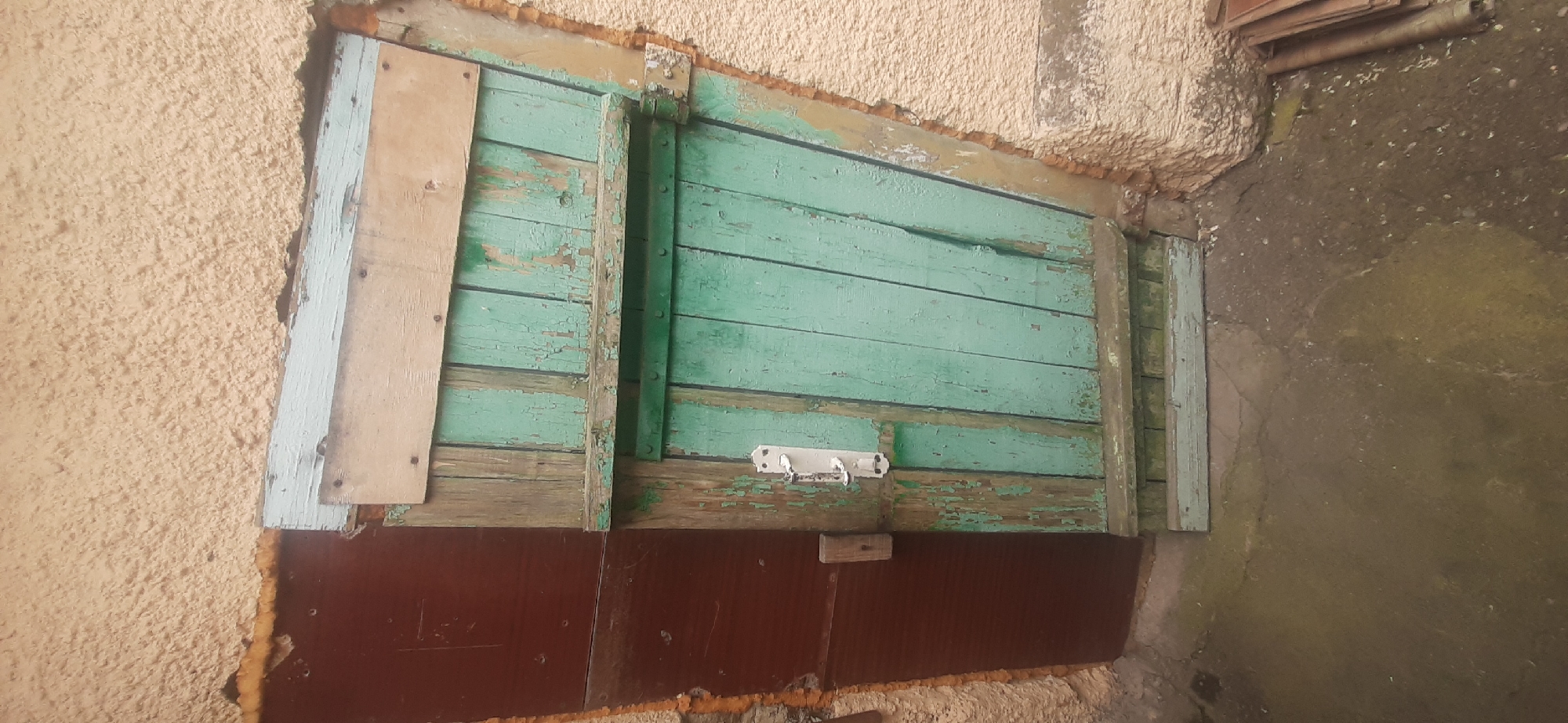 The image shows an old, weathered wooden door, painted green with significant peeling paint, installed in a textured, cream-colored stucco wall. The door is ajar and is partially covered by a dark red door or panel to its left. The door is secured with a white metal latch. Above the green door, there are two horizontal wooden planks. The area around the door opening appears to be rough and possibly insulated with foam. The ground in front of the door is cracked concrete or dirt. To the right of the door, a stack of what appears to be metal roofing sheets is visible. The scene depicts a rustic and somewhat dilapidated entrance. The context of Yessentukskaya, Russia, suggests this might be an exterior door to a shed, outbuilding, or an older residential property. There are no people or animals visible in the image, and no specific actions are taking place.