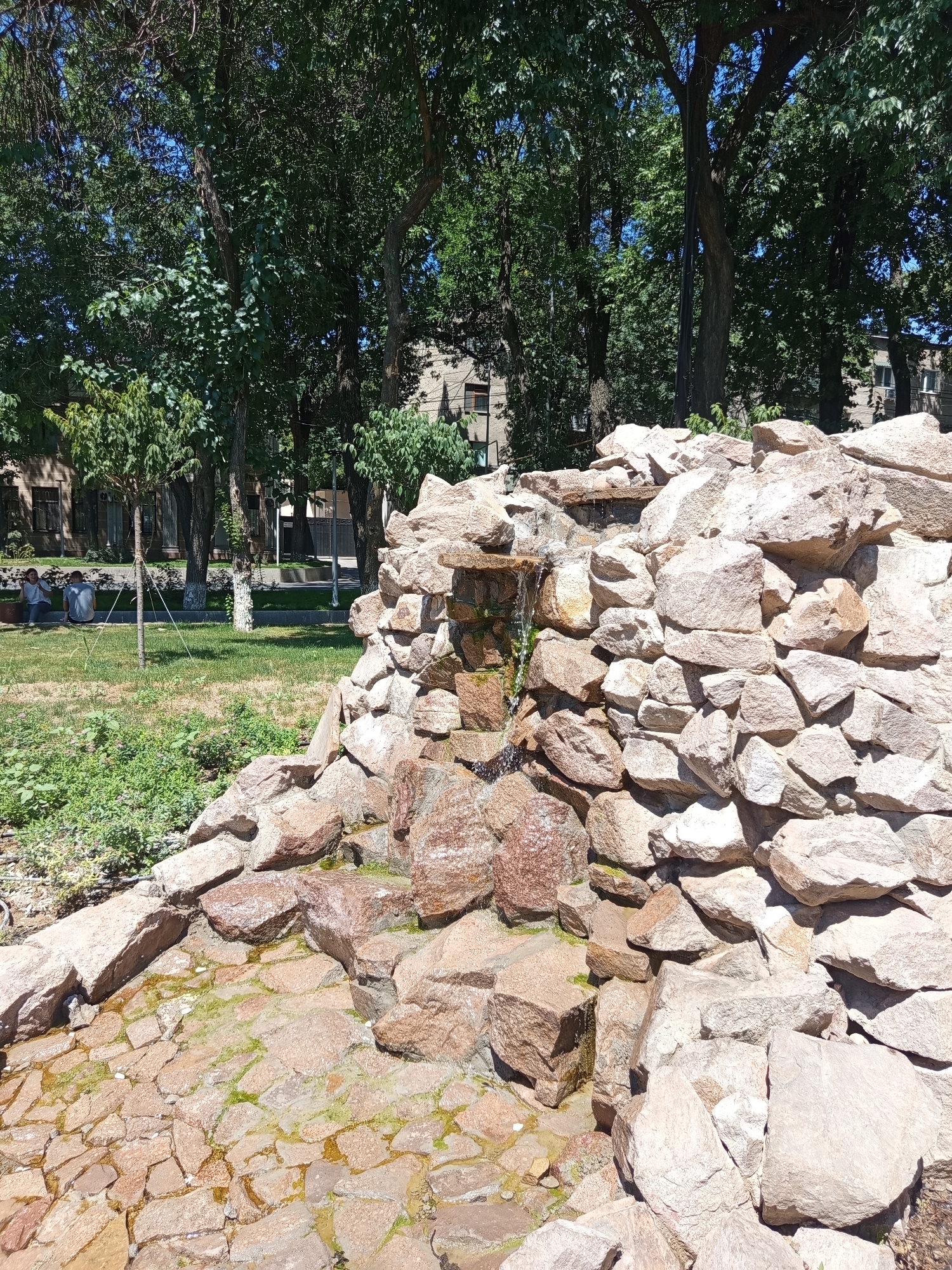 The image shows an outdoor rock formation that appears to be a dry fountain or waterfall feature in a park setting. The rocks are of various sizes and colors, primarily beige and pinkish-red, and are stacked in a way that suggests a natural, unmanicured appearance. Water is visible trickling down some of the rocks and collecting in a shallow basin at the bottom, which is paved with smaller stones. Greenery, including grass and bushes, surrounds the rock feature. In the background, several large trees with full green foliage create a shaded canopy. Behind the trees, a multi-story building with windows is visible, suggesting an urban or suburban environment. Two individuals are seated on a bench in the grassy area in the middle ground, facing away from the camera. The overall scene depicts a tranquil park area, likely within the city of Shymkent, Kazakhstan, on a sunny day.
