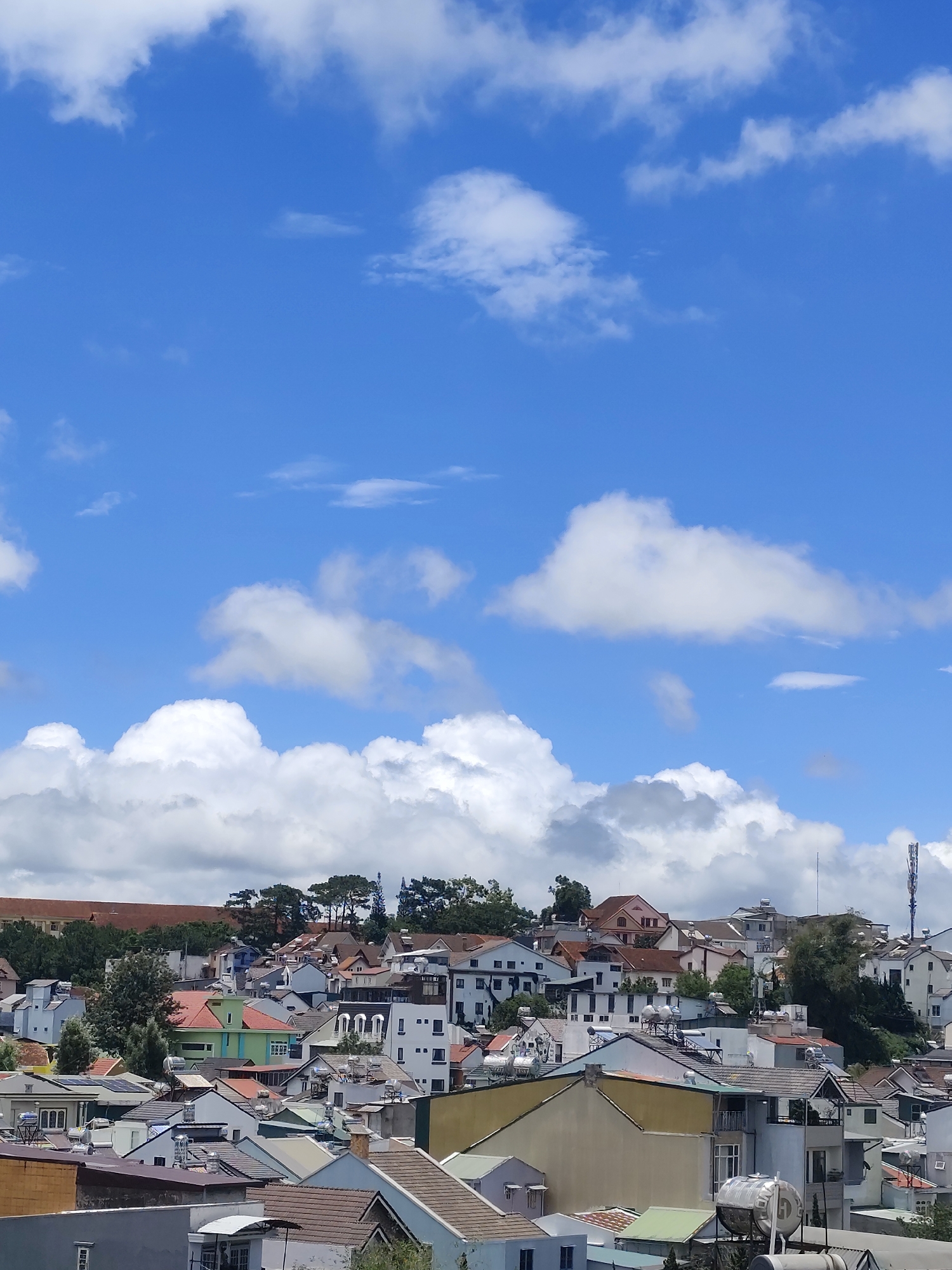 The image shows a cityscape of Da Lat, Vietnam, under a partly cloudy sky. The view is elevated, showcasing a dense arrangement of houses with varied roof colors (red, orange, green, and grey) clustered together. The architecture appears to be a mix of traditional and modern designs, with some houses displaying French colonial influences. Trees are interspersed among the buildings. In the background, a communications tower is visible. The sky occupies a significant portion of the image, featuring white, puffy clouds against a bright blue sky. There are no people visible in the image.
