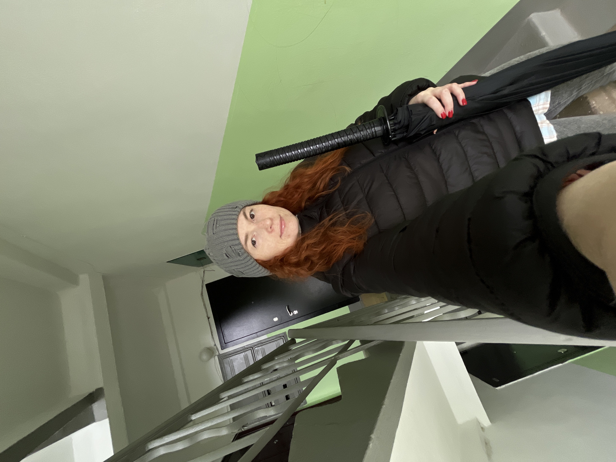 A woman with red hair and a grey beanie, wearing a black puffer jacket, stands on a staircase. She is holding a black umbrella with a katana-style handle. The staircase has white rails and grey steps, and the walls are painted white and green. There is a black electrical box on the wall near the top of the stairs. The location is likely an apartment building. The city is Chaikovsky, Russia.