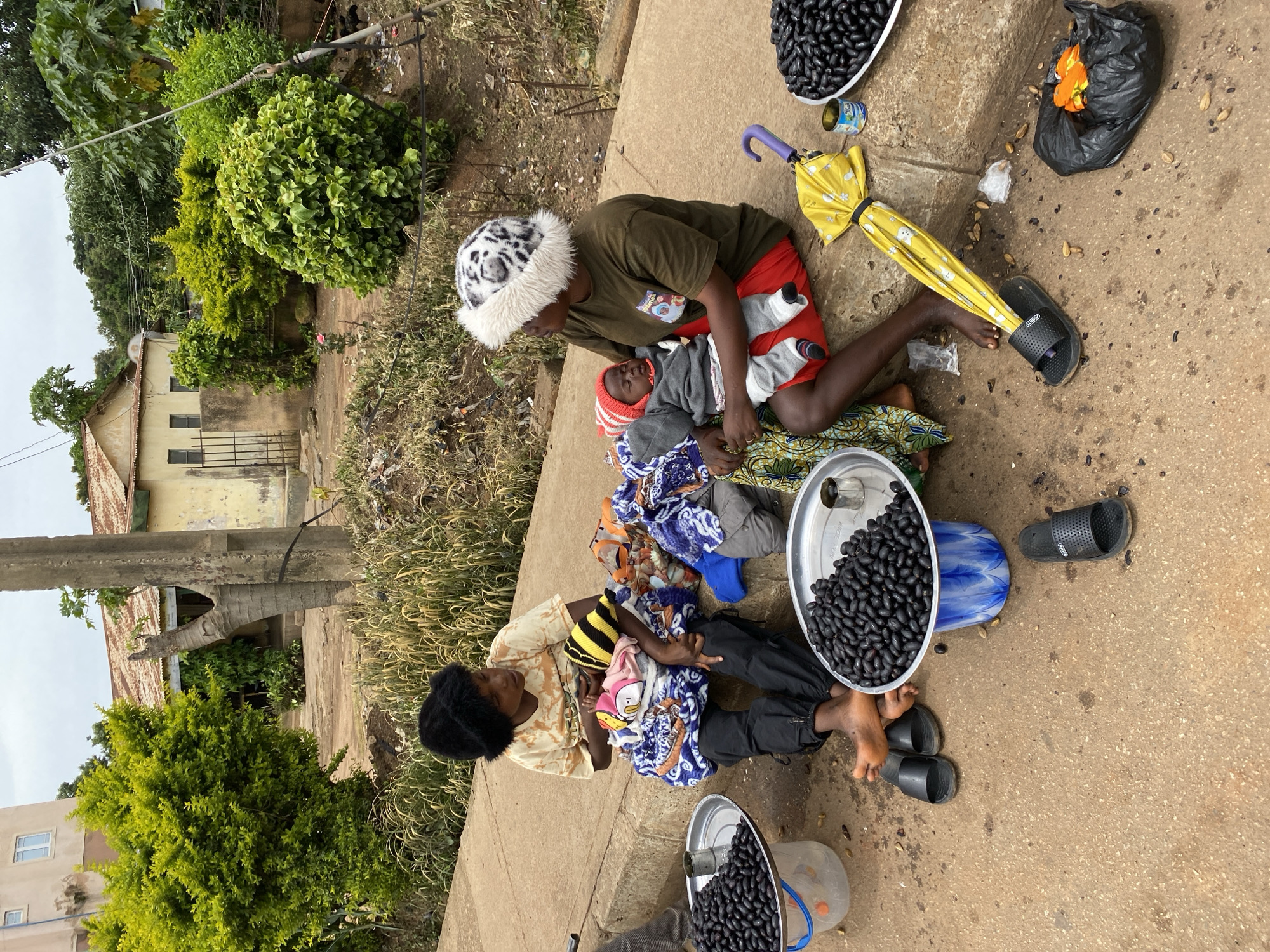 The image shows two women and three babies sitting on a concrete ledge outdoors. One woman, wearing a brown shirt and red shorts, holds a baby wrapped in gray clothes and a red hat. She has a leopard-print hat with white fur. The other woman, wearing a tan shirt and black pants, holds a baby wrapped in colorful blankets and wearing a yellow and black striped hat. A third baby is partially visible, also wrapped in blankets.
In front of the women are several objects. There are two metal trays filled with dark beans, a blue plastic container, and a yellow umbrella with purple accents. A black trash bag with an orange object inside is also present, as are two pairs of black flip-flops scattered on the ground. The ground is a mixture of concrete and dirt, with some scattered beans and small objects.
The scene appears to be a roadside or market area. In the background, there is a building with a weathered facade and a corrugated metal roof. Lush green trees and foliage are visible, suggesting a tropical or subtropical environment. The sky is overcast, indicating a cloudy day. The overall mood is one of quiet activity, possibly a vendor selling beans. There is no visible text in the image. The location is identified as Bukuru, Nigeria.