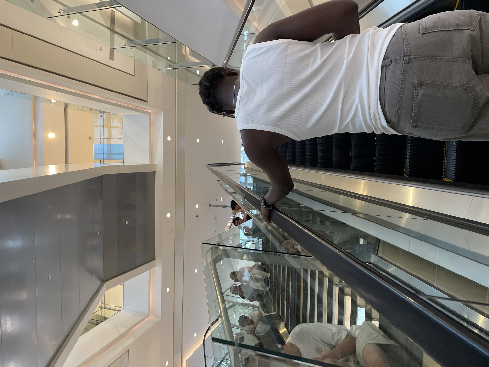 In a multi-level indoor environment, likely a modern shopping mall in Yuzhong District, China, a Black person is ascending an escalator. The individual is wearing a white ribbed tank top and grey jeans, with their back to the camera. They are holding onto the escalator's handrail, with a watch visible on their left wrist. To the left of the escalator, a glass-paneled staircase is visible, occupied by several individuals seated on the steps, many looking at their phones. The overall scene depicts a busy public space with architectural elements such as glass railings, polished floors, and modern lighting.