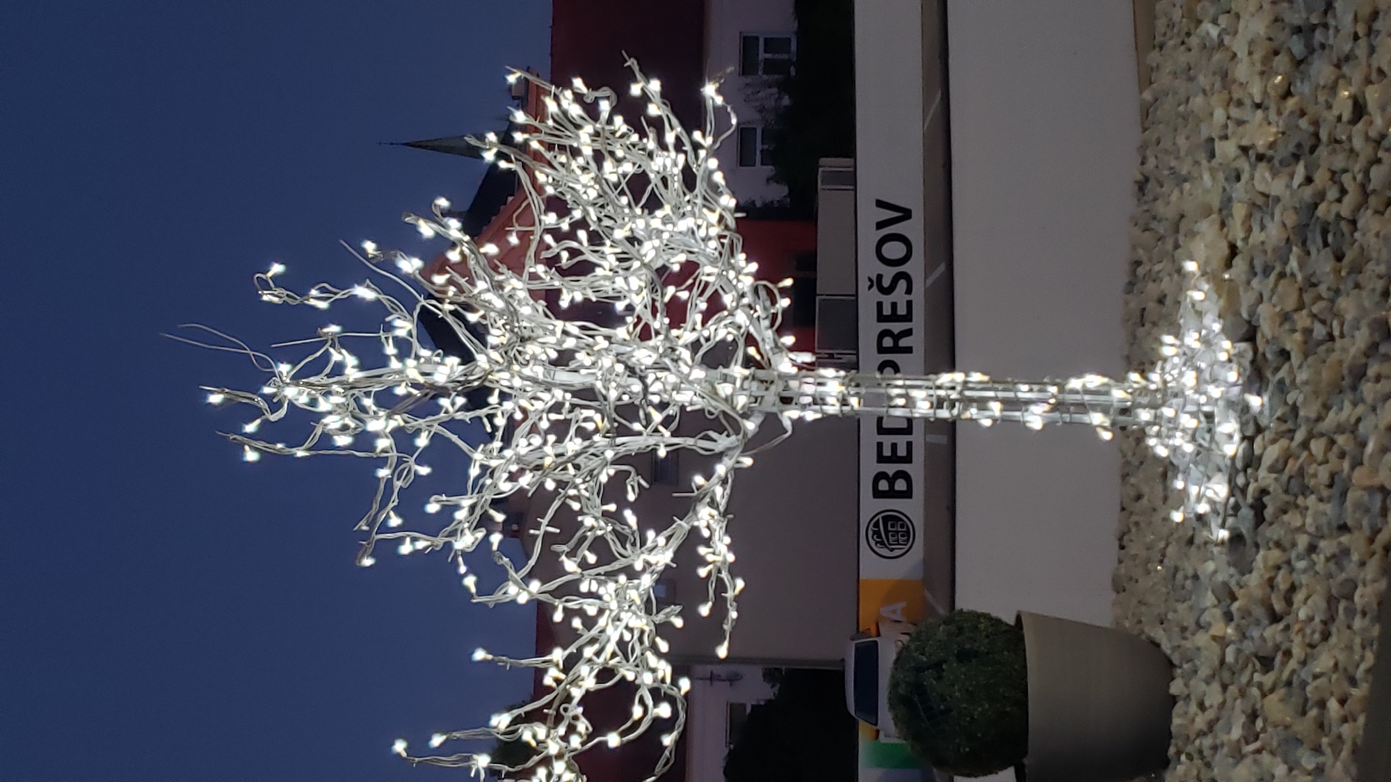The image shows an illuminated artificial tree with white LED lights, situated outdoors at dusk. The tree is adorned with numerous small white lights that are wrapped around its branches and trunk. In the background, there are buildings with red roofs and a spire, suggesting an urban or town setting. A white banner with dark lettering is visible behind the tree, which reads "BEDPREŠOV". To the left of the tree, a potted plant with a spherical green top is partially visible. The ground is covered in light-colored gravel. The sky is a deep blue, indicating twilight or evening. The overall scene suggests an outdoor decoration, possibly for an event or as a permanent fixture in a public space in the city of Prešov, Slovakia.
