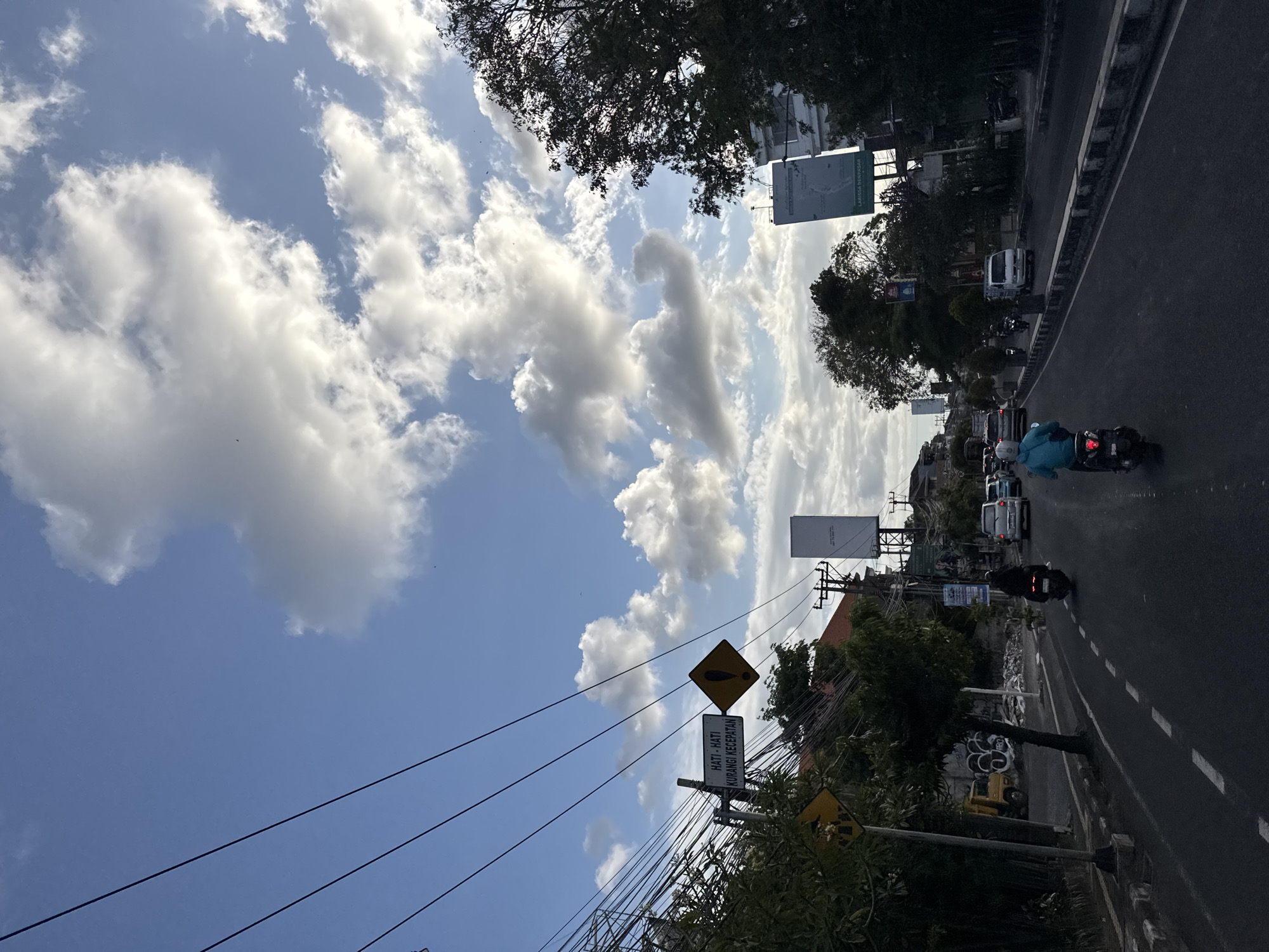 The image captures a street scene in Denpasar, Indonesia, under a partly cloudy sky. The perspective is from a vehicle traveling on a road. Several utility wires crisscross the sky. On the left side of the road, a yellow diamond-shaped warning sign with a black border and exclamation mark is mounted on a pole. Below it, a white rectangular sign reads "HATI-HATI KURANGI KECEDATAN" which translates to "CAUTION, REDUCE SPEED." Further down the road, traffic includes motorcycles and cars. One motorcycle rider in a blue jacket is prominent in the foreground, traveling in the same direction. On the right side of the road, tall trees and buildings line the street, with billboards visible. A yellow truck is parked on the left side, near a wall with graffiti. The road is paved with dashed white lines marking the lanes. The overall impression is a typical urban street environment in Indonesia during the daytime.
