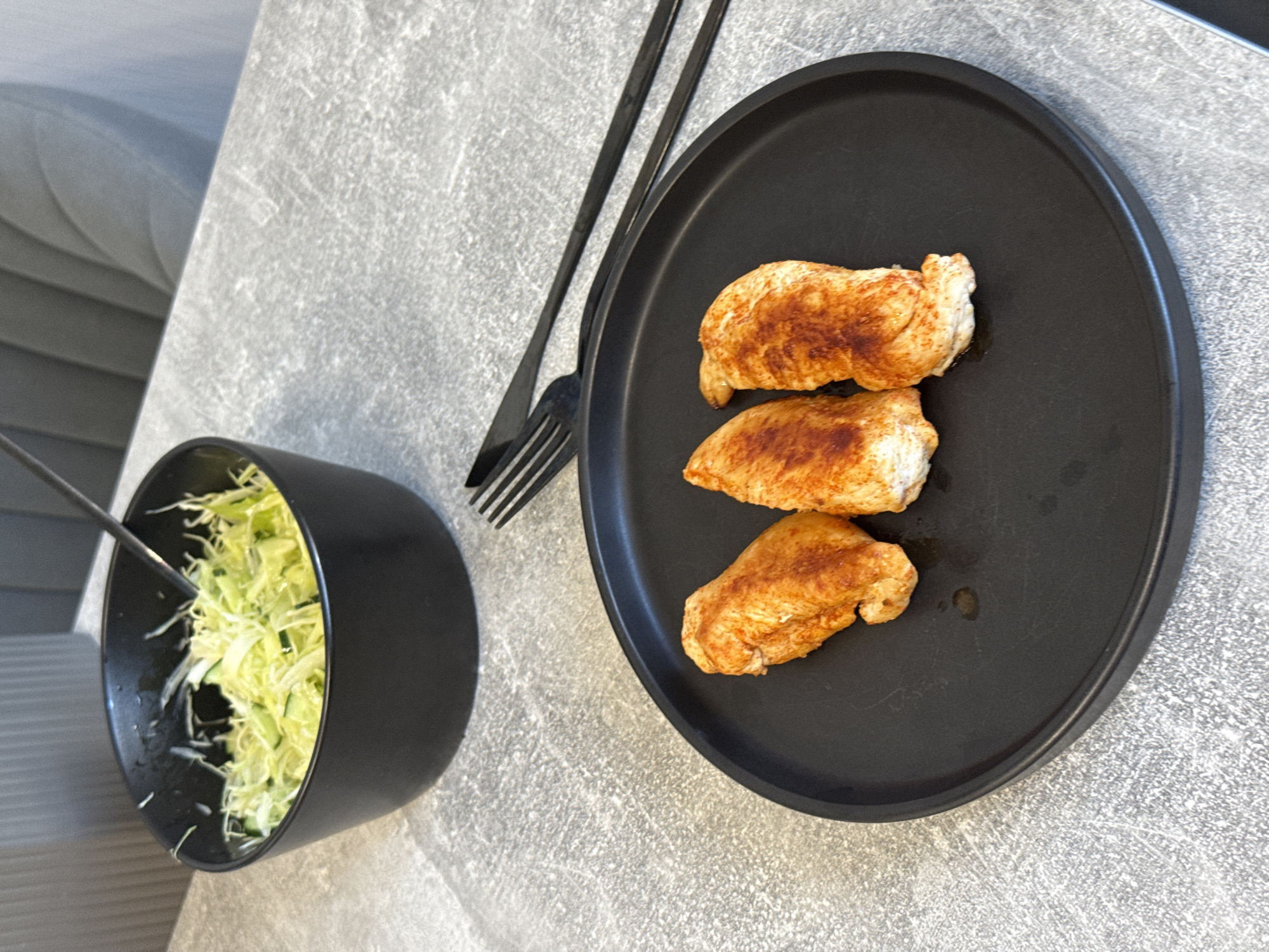 A black plate with three pieces of seasoned chicken breast sits on a gray, textured surface. Beside the plate is a black fork and knife set. In the background, a black bowl filled with shredded cabbage and sliced cucumber is visible. The scene appears to be set in a dining area, possibly in Lyubertsy, Russia, though no specific landmarks or identifying features confirm this location. The lighting is natural, casting soft shadows.