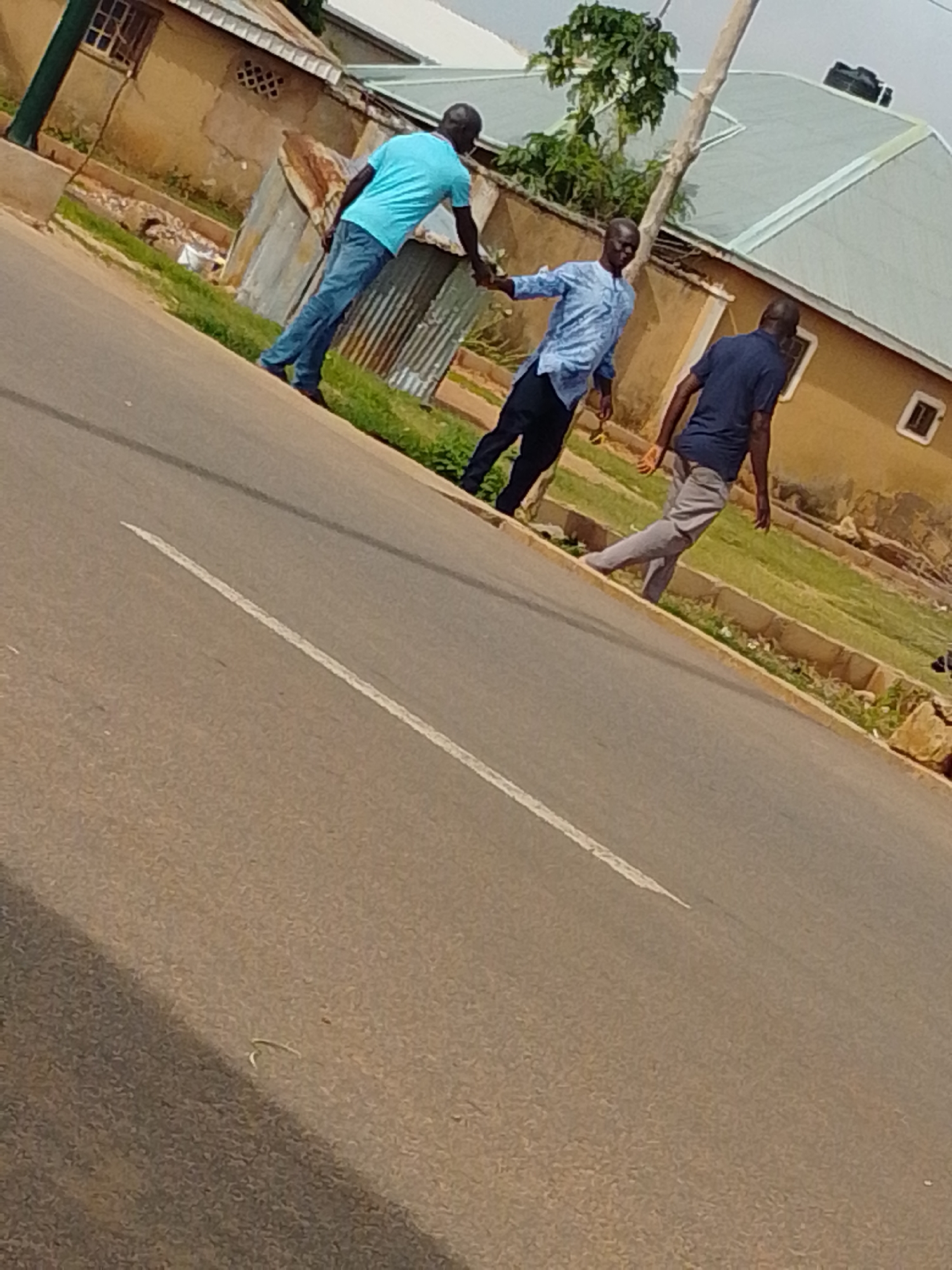 The image depicts three men on a street in Gombe, Nigeria. The street is paved with asphalt and has a white line marking. The men are walking and interacting with each other. The man on the left is wearing a blue t-shirt and jeans. The man in the middle is wearing a light blue patterned shirt and dark pants. The man on the right is wearing a dark blue t-shirt and light gray pants. They appear to be holding hands or reaching for each other's hands. In the background, there are buildings with corrugated metal roofs and beige walls, as well as some vegetation and a utility pole. The overall scene suggests a casual outdoor interaction in a semi-urban environment.