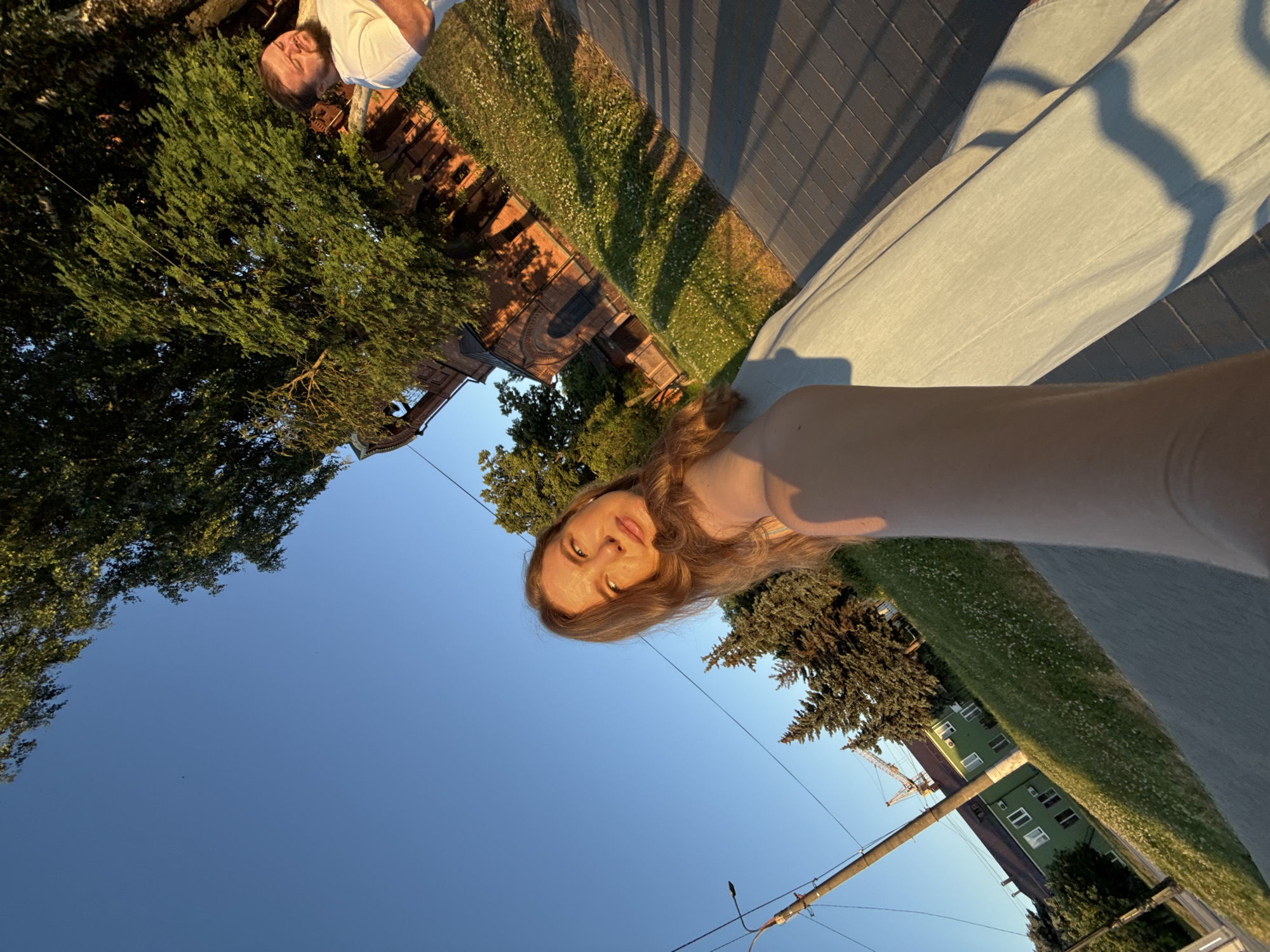 A low-angle, wide-shot photograph captures a sunny day scene, possibly on a street or pathway in Vladimir, Russia. In the foreground, a young woman with long, blonde hair is partially visible, her arm extended towards the camera as if taking a selfie. She is wearing a light-colored dress and is looking upwards and to her right, with sunlight highlighting her face.
In the mid-ground, a man with a beard and light-colored t-shirt is visible, looking to his left with his eyes closed, seemingly enjoying the sun. Behind him, a red brick building with arched windows, possibly a church or historic structure, is partially obscured by lush green trees. A pathway with distinct shadow patterns, likely from the trees, runs diagonally across the frame.
The background features a clear blue sky, interspersed with the dark silhouettes of trees and a street lamp on the left side of the image. Further in the distance, a green building with white-framed windows is visible, and a construction crane can be seen over the rooftops, suggesting ongoing development in the area. The ground is covered with grass dotted with small white flowers. The overall lighting indicates late afternoon or early evening sun.