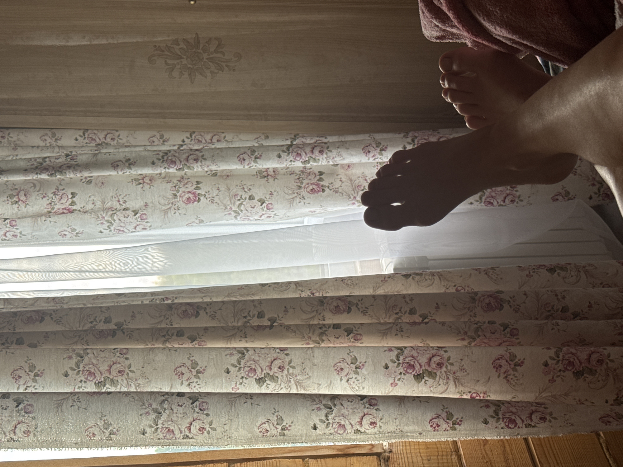 The image shows a close-up view of a person's feet, which are resting on a pink blanket. The feet are in the foreground, and the background is out of focus. The background appears to be a window with floral curtains and a wooden wall. The lighting suggests that it is daytime. There is no indication of the specific location within Nikolaevka, Russia, or any specific actions or interactions occurring. The scene is presented in a static and observational manner.