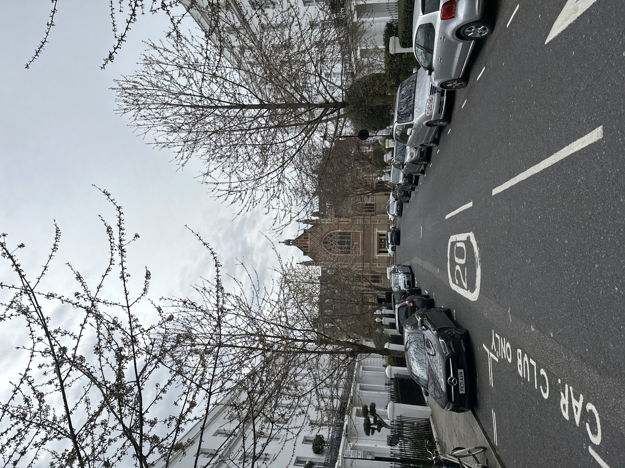 The image captures a street scene in London, United Kingdom, featuring a row of parked cars lining the side of the road. The road is marked with "CAR CLUB ONLY" and a "20" speed limit sign. Bare trees stand on both sides of the street, framing the view of a distant building with distinctive gothic-style architecture. The sky is overcast. White buildings and black iron fences line the left side of the street.