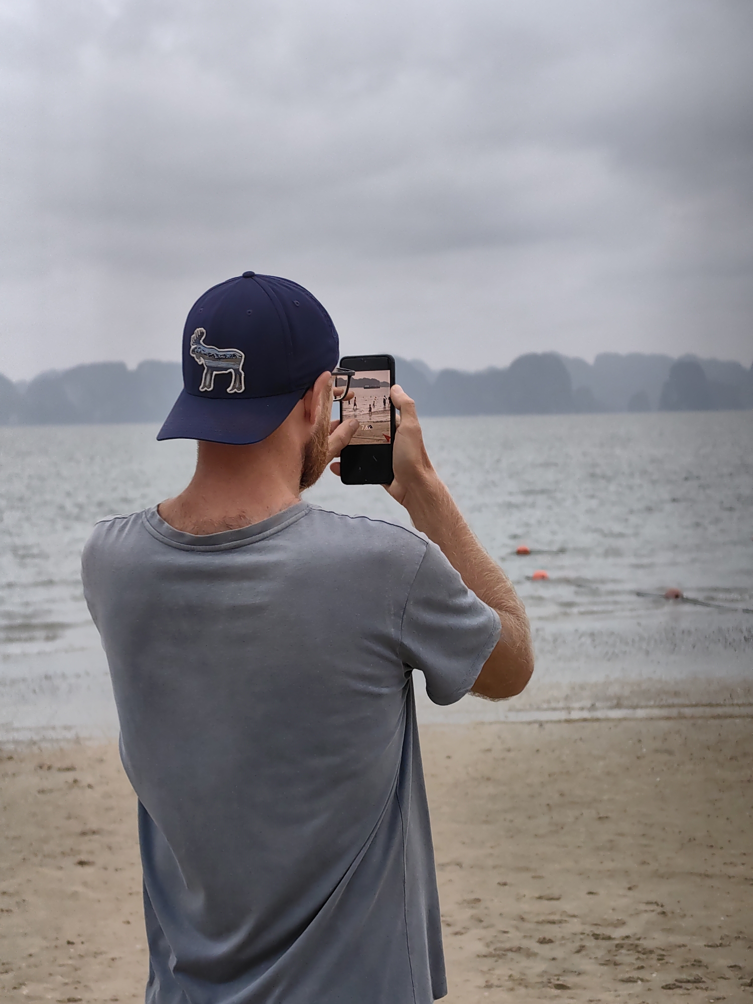 The image features a man, viewed from behind, standing on a sandy beach and actively taking a photograph with his smartphone. He is positioned facing a large body of water, with distinct landmasses visible in the distance.
The man is wearing a navy blue baseball cap that has a white and blue embroidered logo of a moose or elk on its front. He also wears a plain light grey short-sleeved t-shirt. A short, light-colored beard is visible on the side of his face and neck. His arms are raised to hold a black smartphone in a vertical position, and the screen clearly displays an image of several people on a beach.
The setting is a tranquil beach scene under an overcast sky. The sky is a uniform light grey, suggesting a cloudy day. The sea is calm, with gentle ripples on its surface, and a few red buoys are scattered in the mid-distance. The horizon is characterized by numerous dark, irregular shapes of limestone karsts or islands, typical of the landscape found in Halong Bay. The foreground shows a light brown sandy beach, appearing wet near the water's edge. The location is Bai Chay Ward, Vietnam. The time of day appears to be daytime, though the cloud cover subdues the light. No other distinct text is visible apart from the stylized animal logo on the cap.