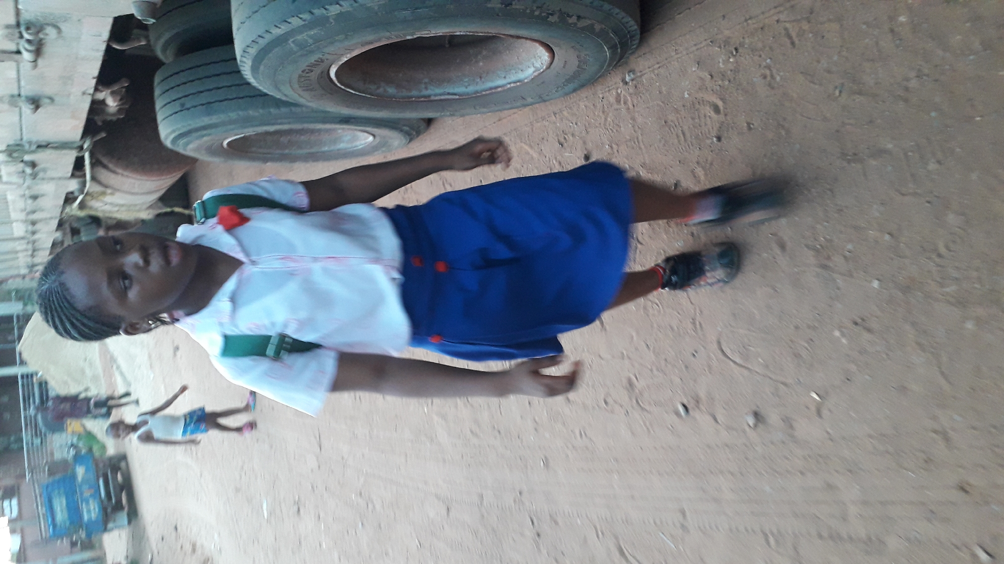 A girl in a white and blue school uniform is walking on a dirt road in Nkpor, Nigeria. She has a green backpack. A truck with large tires is parked nearby. In the background, other people are visible, and there is a blue vehicle. The ground is dirt and dust.