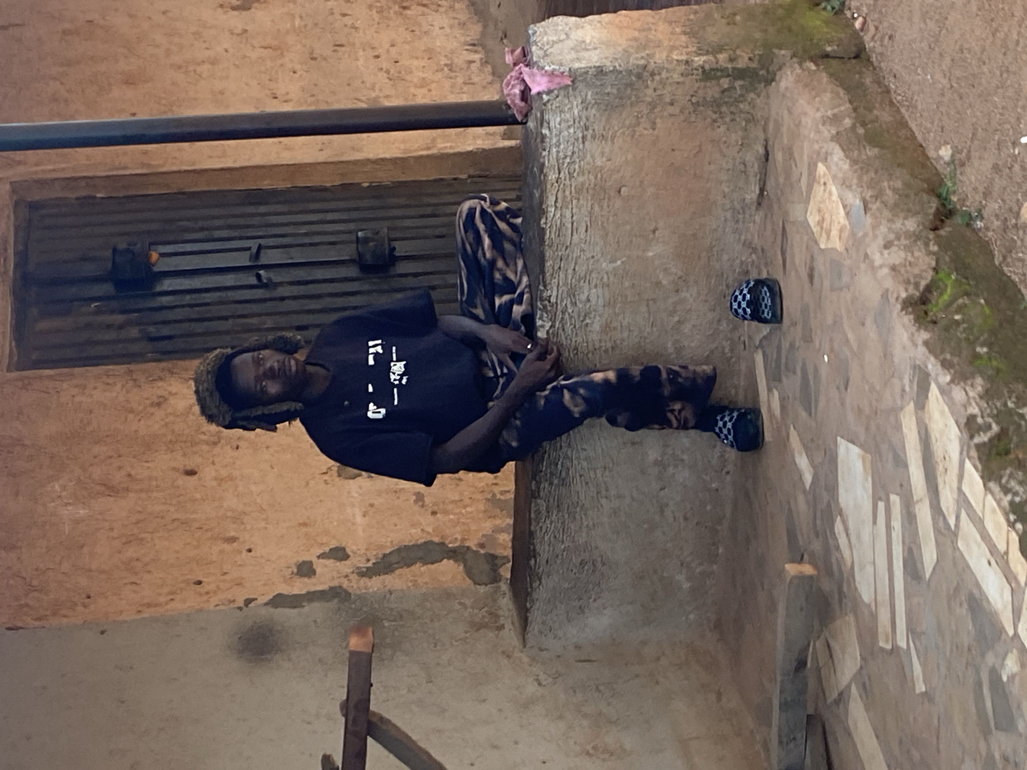 A young Black male with dreadlocks and a fuzzy hat sits on a raised concrete ledge in front of a textured, earthy-brown building with a dark wooden door. He wears a black t-shirt with white lettering and patterned pants. His feet are bare except for black and white checkered slides. To his left, a wooden beam is attached to the wall. The ground in front of the ledge is paved with irregular light-colored stones and some scattered wooden pieces. A pink cloth is draped over the ledge to the right of the man. The overall scene suggests an outdoor, possibly informal setting in Bukuru, Nigeria.