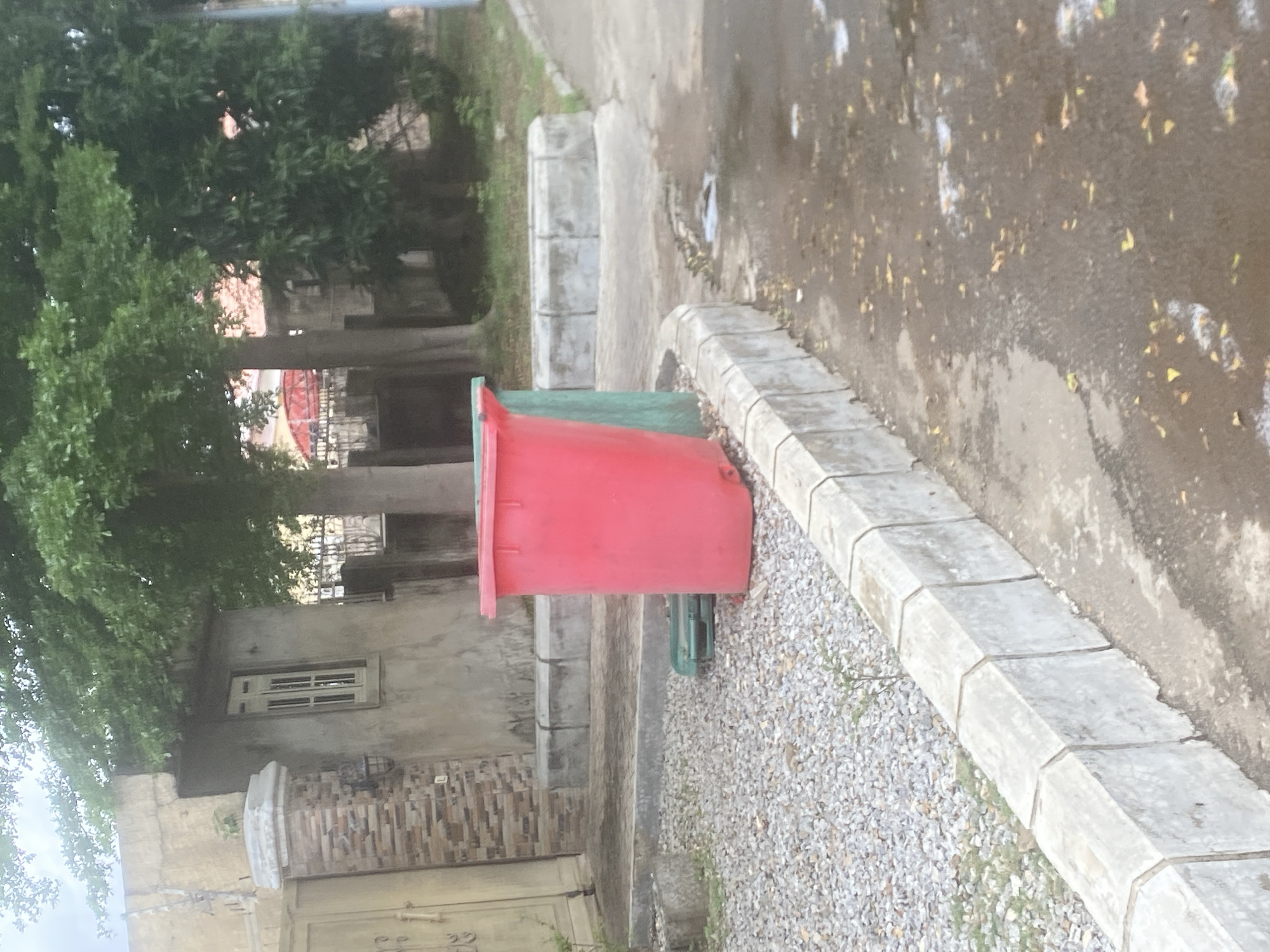 A red trash bin and a green recycling bin are situated on a gravel-covered area next to a concrete curb in Gosa, Nigeria. Behind the bins, a portion of a building is visible, featuring a tan-colored facade and a window. A tall tree with lush green foliage is prominent in the background, casting shadows on the scene. The ground in front of the curb is paved and appears wet, with fallen leaves scattered across it.