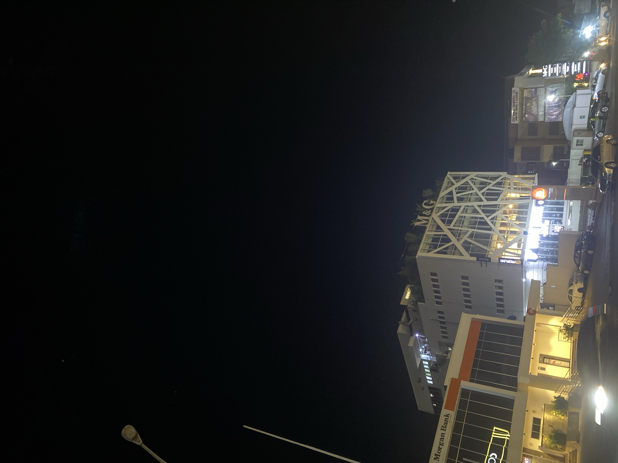 The image shows a night scene in Abuja, Nigeria. The foreground features a lit commercial building with "Morgan Bank" signage. Adjacent is a taller white building with a geometric steel structure on top, bearing the letters "M&G". Cars and streetlights are visible on the right, suggesting a road or street. In the background, additional buildings and illuminated signs are visible. The sky is dark and clear.