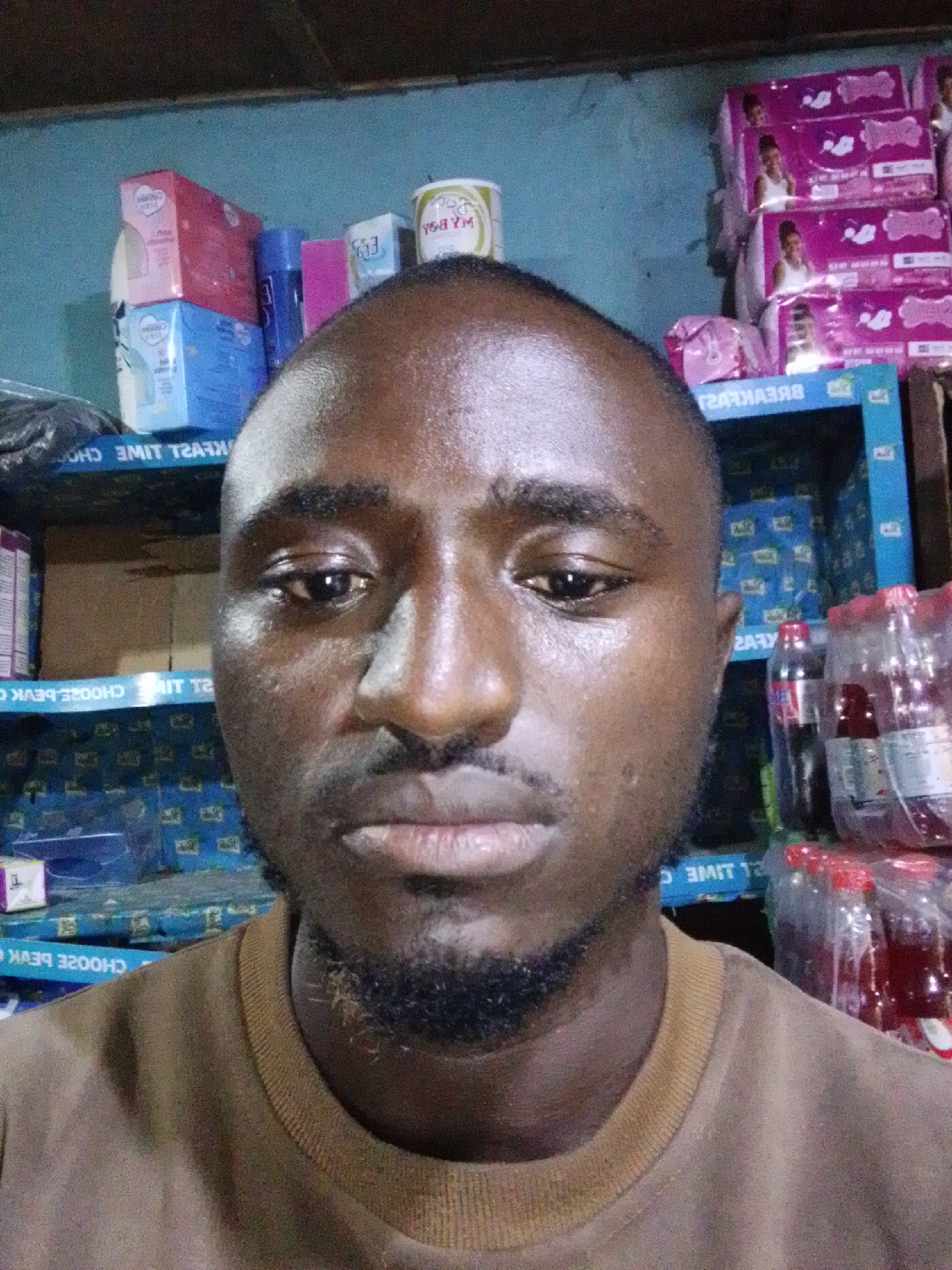 Here is a detailed description of the image:
**Subject:** A close-up portrait of a dark-skinned man with a closely shaved head, short beard, and mustache. He appears to be indoors.
**Setting/Environment:** The background seems to be a small shop or store, evidenced by shelves stocked with a variety of products. Items include toiletries, sanitary products (like feminine pads stacked up), and beverages (like bottled drinks). The shelving unit behind the subject is a light blue color. There are also other indistinct items further back on the shelves.
**Objects:**
* Various packaged goods are visible on shelves, like tissues or toilet paper.
* Bottled beverages are stacked to the right of the subject.
* Shelving unit holding the products.
* The man is wearing a brown t-shirt.
**People:**
* The primary subject is the man in the foreground. No other individuals are clearly visible.
**Action/Interaction:** The man is looking directly at the camera, implying the photo was taken by someone or something he is facing.
**Location:** This picture was taken in the city of Unknown, Unknown.