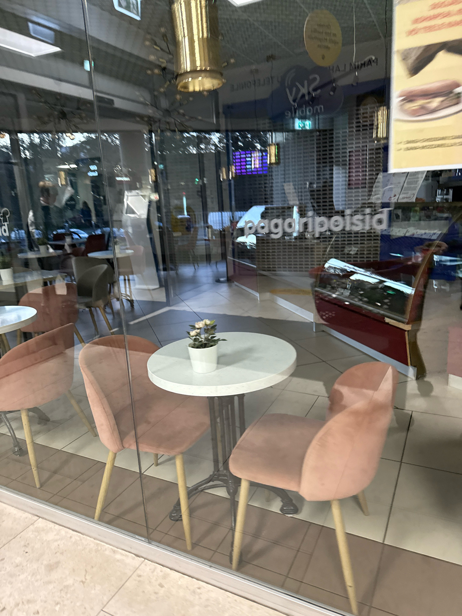 The image shows the interior of a cafe as seen through a glass window. The cafe appears to be located in Tallinn, Estonia, as indicated by text on the window. Inside, there are several tables and chairs, with some chairs being a light pink color. A small white table with a white pot containing a flowering plant is visible in the foreground. In the background, there are more tables and chairs, a red display case, and shutters on the windows of other establishments. Reflections on the glass include trees and buildings. The overall impression is that of a modern cafe interior, possibly in a shopping mall or similar commercial area.